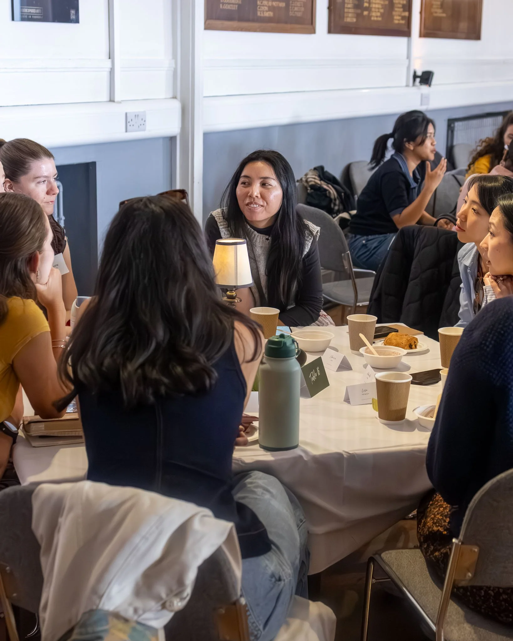People sitting around a table at a casual gathering or meeting, engaging in conversation. The table has food and drinks, and a lamp is visible in the center. The background features framed boards on the wall.