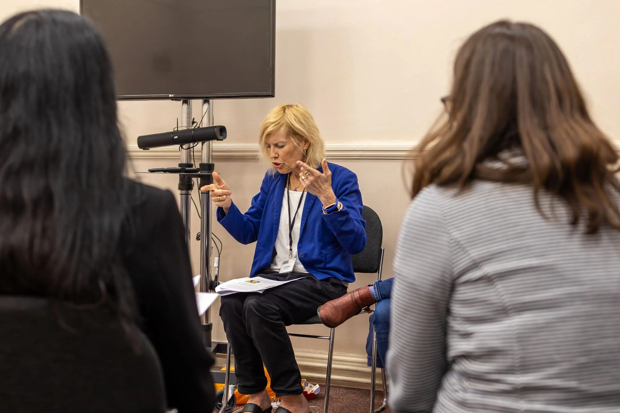 A woman with blonde hair wearing a blue blazer and white shirt sitting in front of a large television screen, surrounded by three people, two women and one man, who are facing her.
