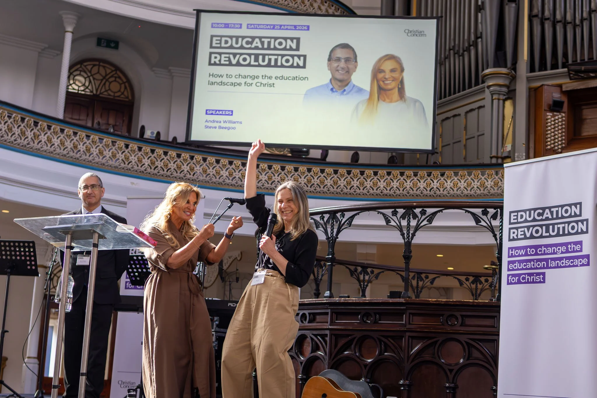 People on stage at a conference with a large screen displaying a presentation about an education revolution, date April 25, 2026, featuring two speakers, Andrea Williams and Steve Beegoo, in a grand hall with ornate railing and organ pipes in the bac