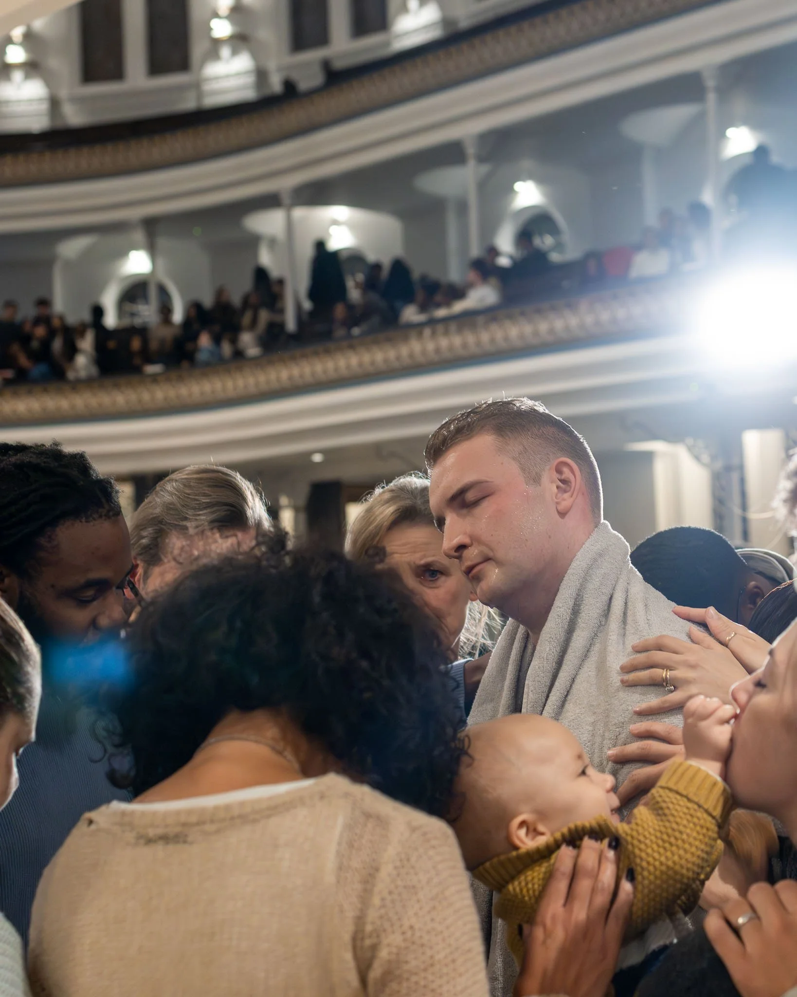 Group of people praying closely together in an indoor setting with multiple balcony levels and ornate architecture.