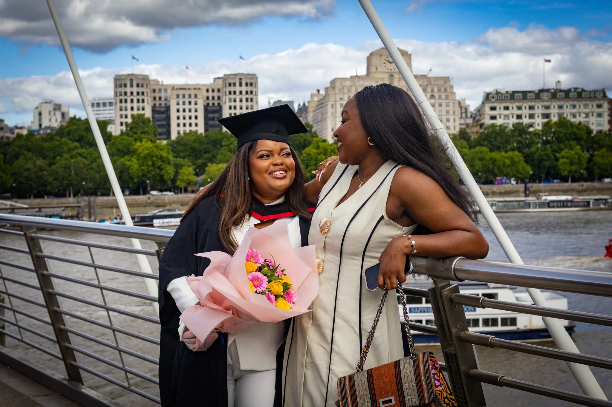 Two women share a joyful moment on a bridge in London against a cityscape background. One woman, in a graduation cap and gown, holds a bouquet of pink and yellow flowers. The other woman, dressed in a sleeveless striped jumpsuit, hugs her warmly.