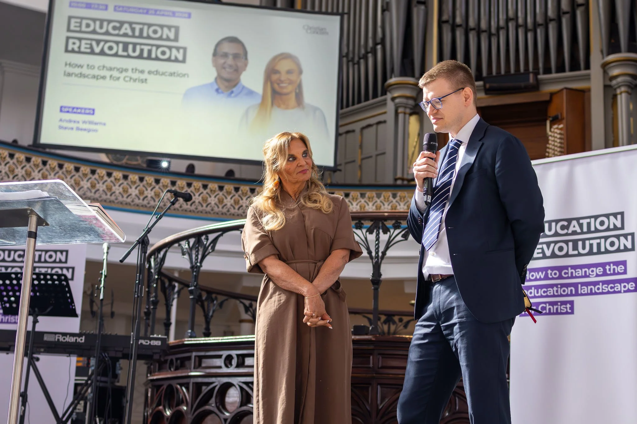A man in a suit speaking into a microphone and a woman in a brown dress listening, in front of a large screen displaying a presentation titled "Education Revolution" at a church or auditorium.