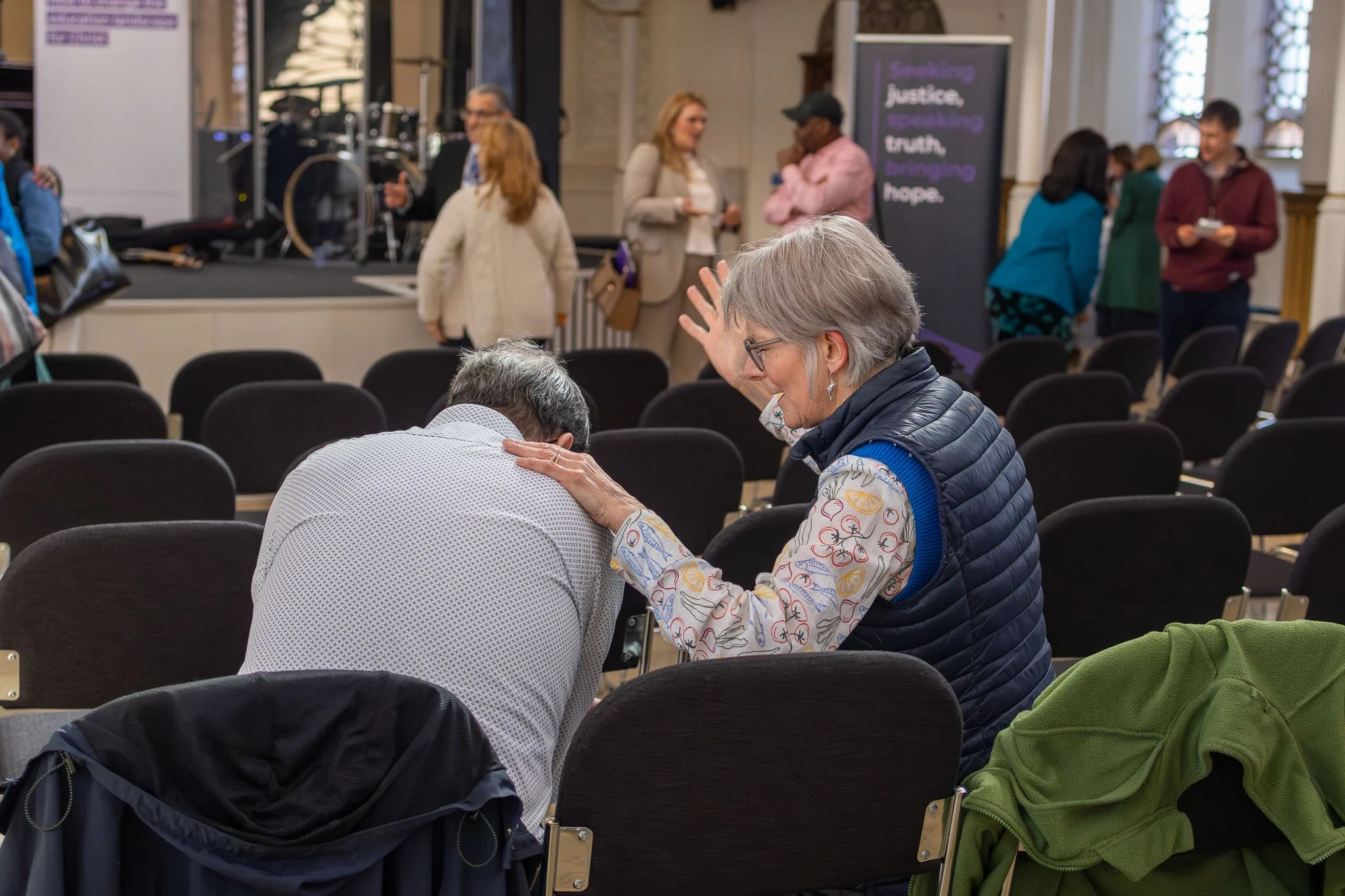 People praying and connecting with each other in a church, with a focus on a woman with gray hair and glasses gently praying over a man