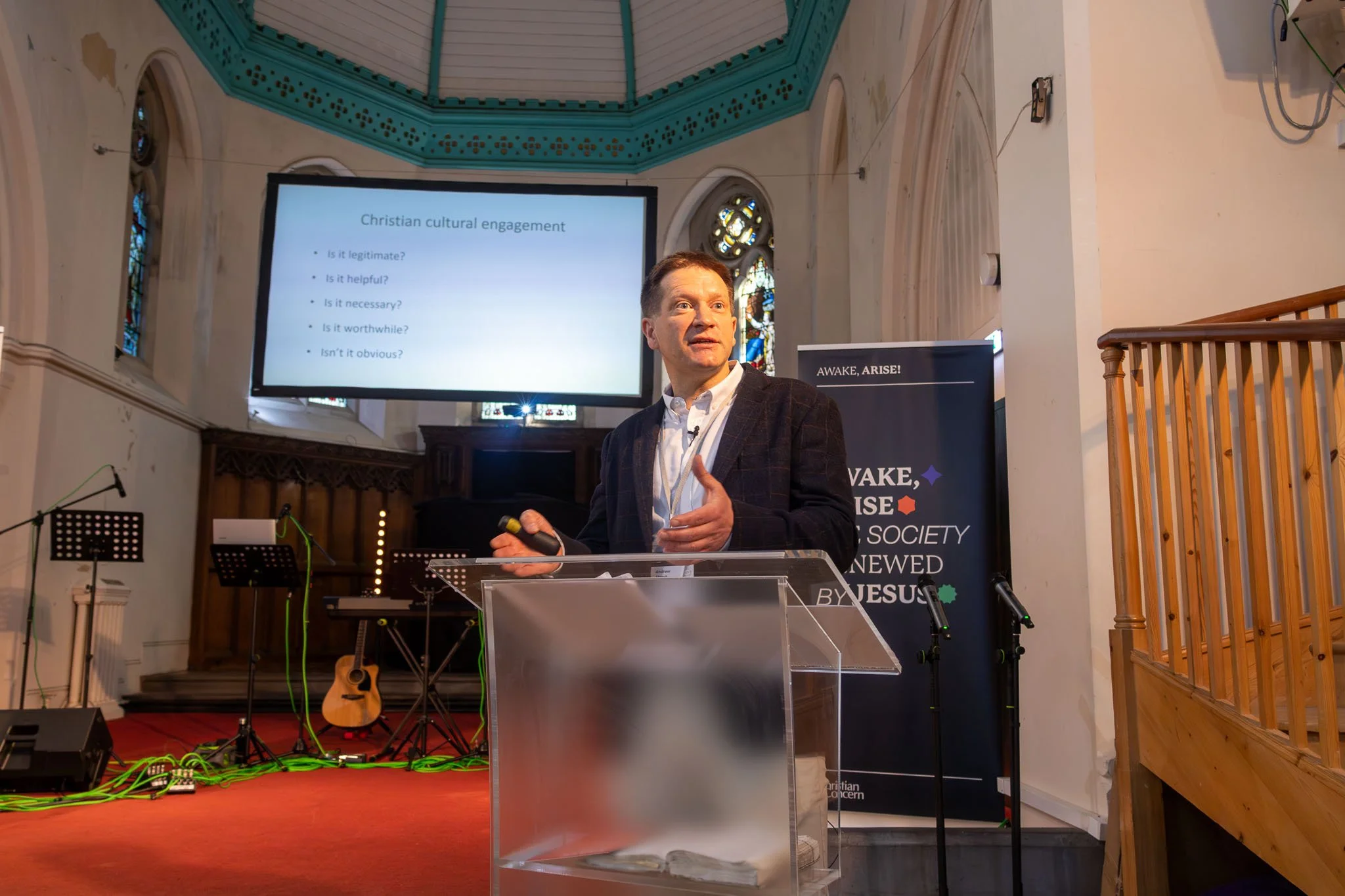 A man in a dark blazer and white shirt giving a presentation in a church-like setting. A large screen behind him displays the title 'Christian cultural engagement' with several questions listed underneath. There are stained glass windows behind the s