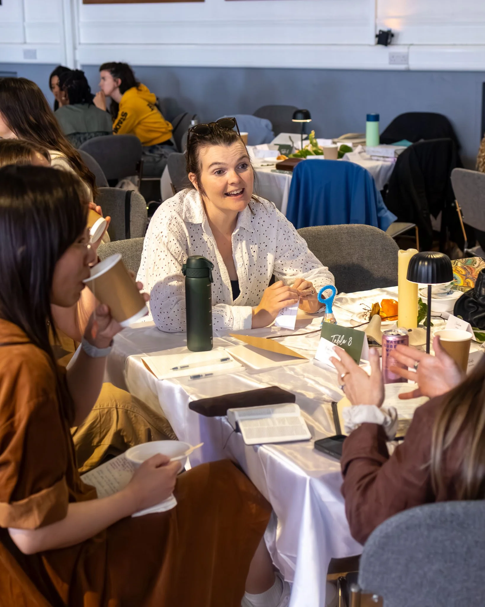 People sitting at a table having a discussion, some holding cups, with notes and books in front of them.