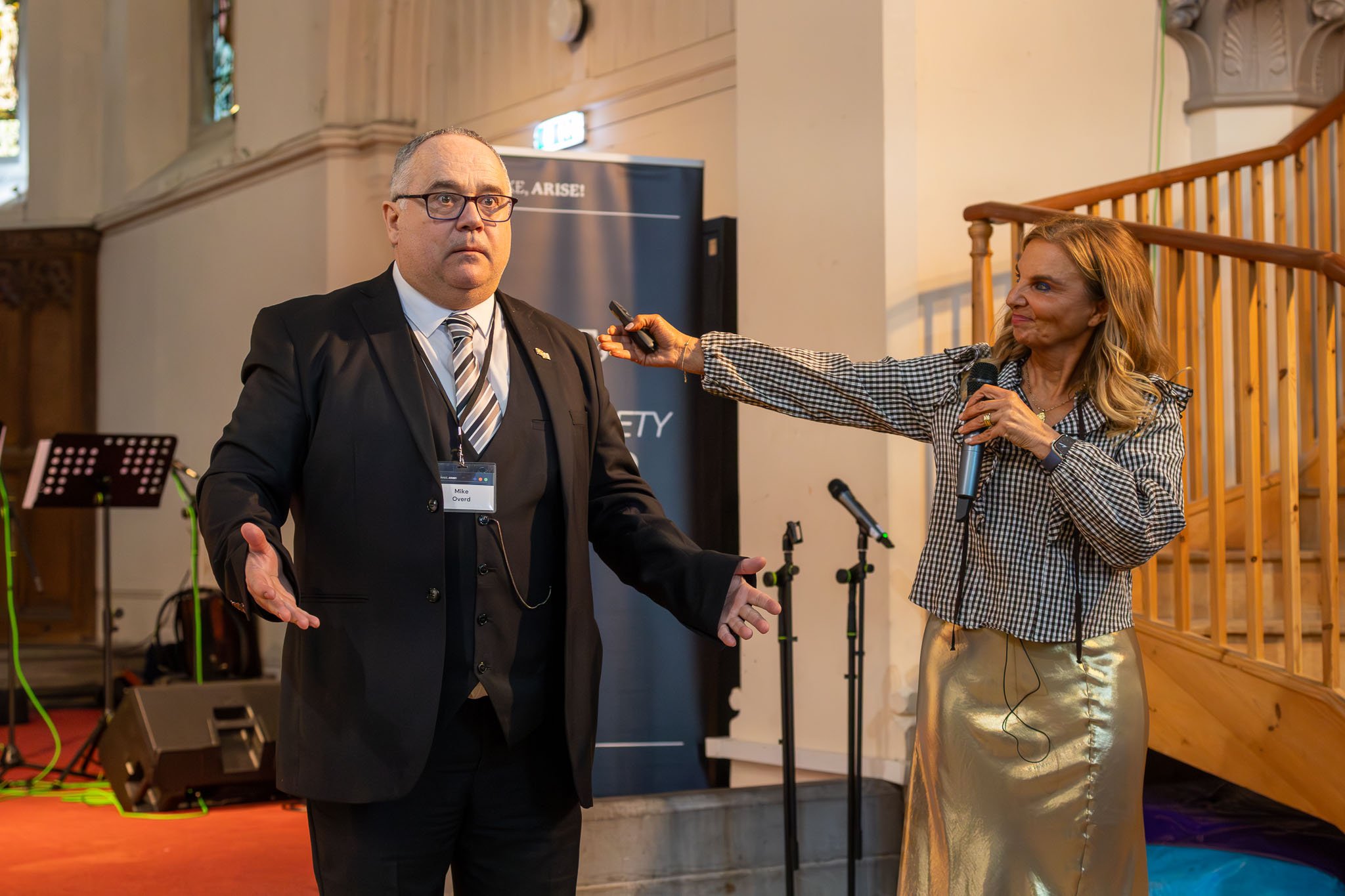 A man in a black suit and glasses stands with his arms outstretched, speaking to an audience. A woman holding a microphone is pointing a recording device towards him. The setting is a church or hall with a wooden railing, musical equipment, and a ban
