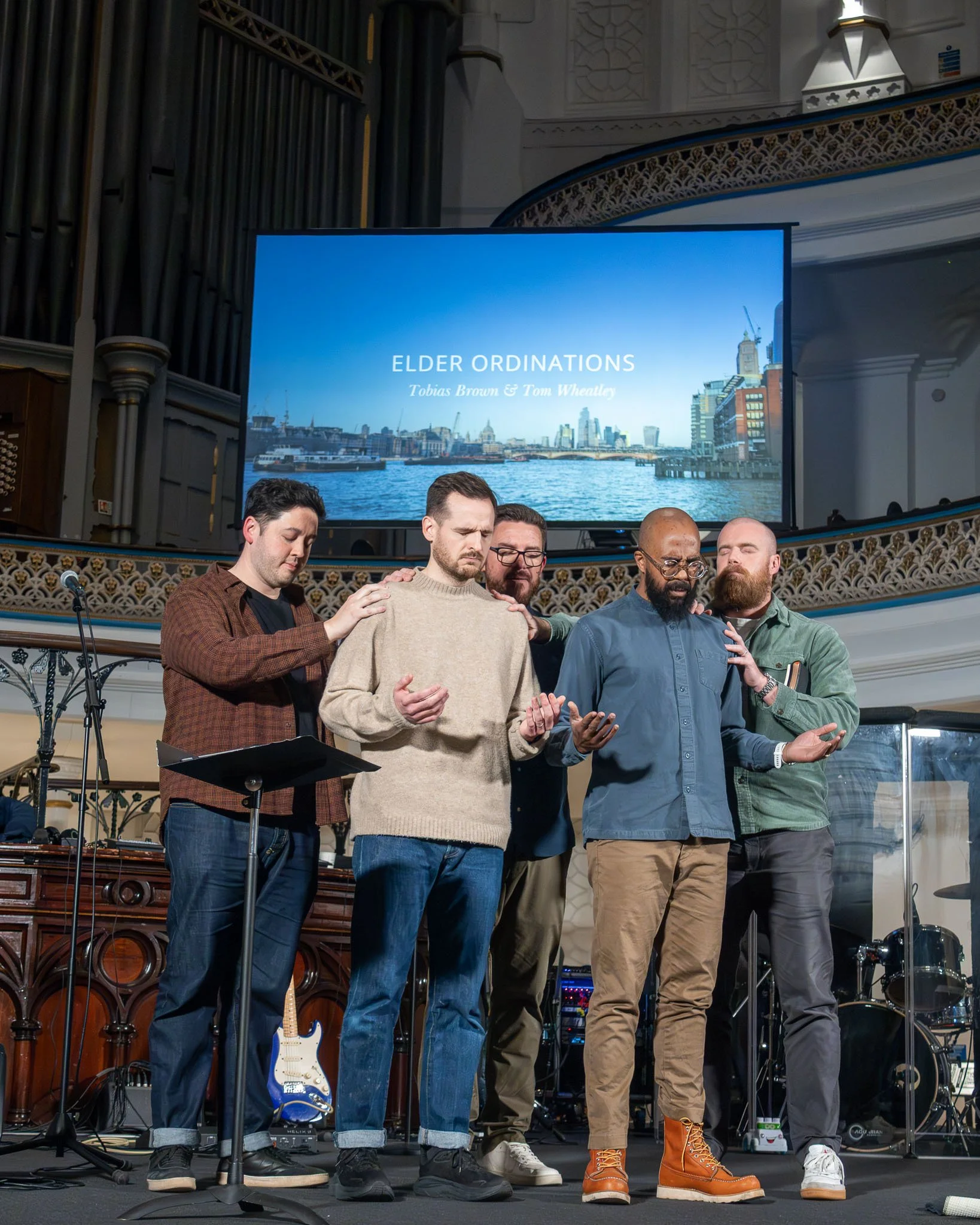 A group of men praying with their eyes closed, standing with hands on each other's shoulders, inside a church. Behind them, a large screen displays the text 'Elder Ordinations' with names Tobias Brown and Tom Wheatley, along with a cityscape backgrou