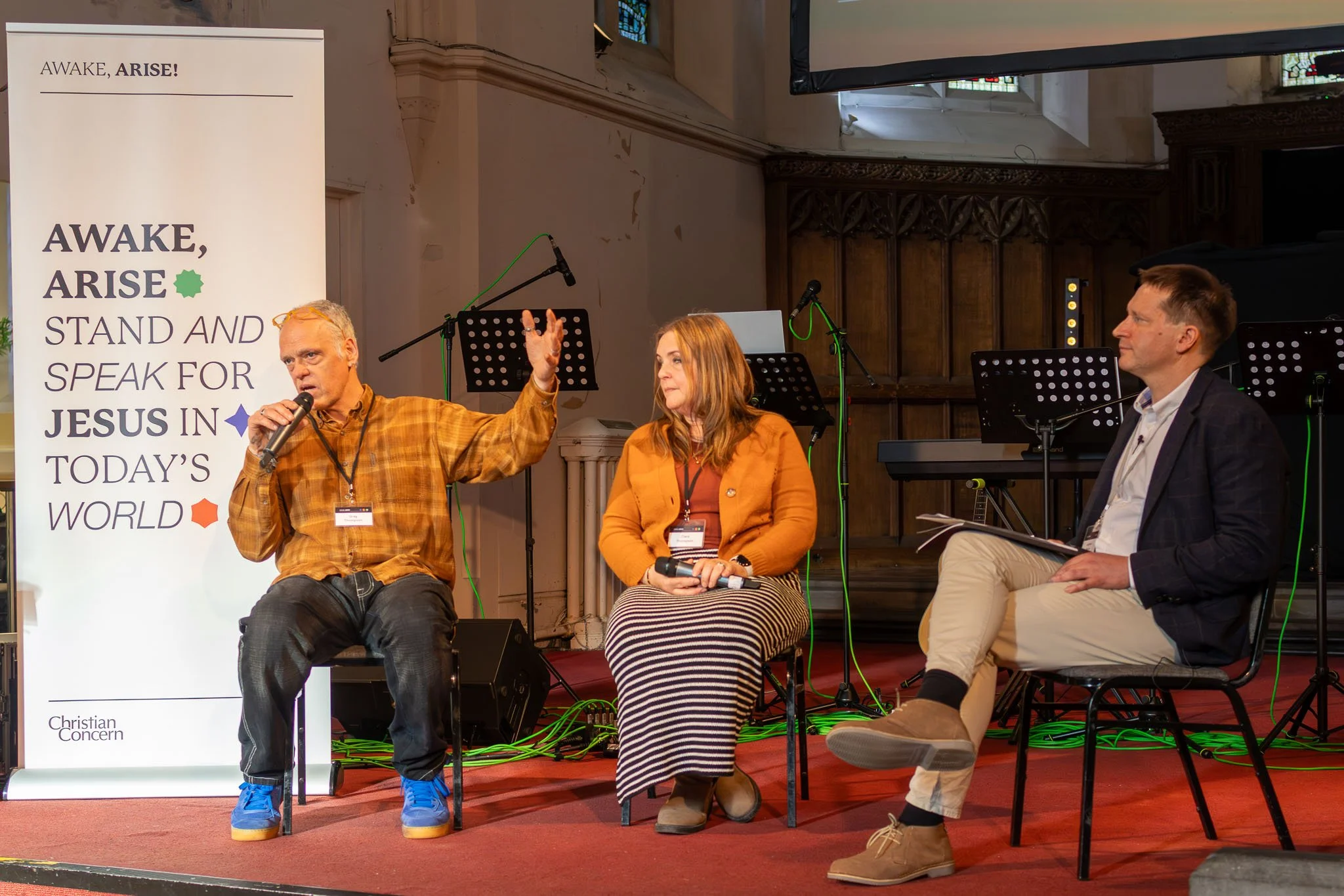 Three people seated on a stage during a discussion, with a man on the left speaking into a microphone, a woman in the middle, and a man on the right holding a notebook with a light background and a religious-themed banner to their left.