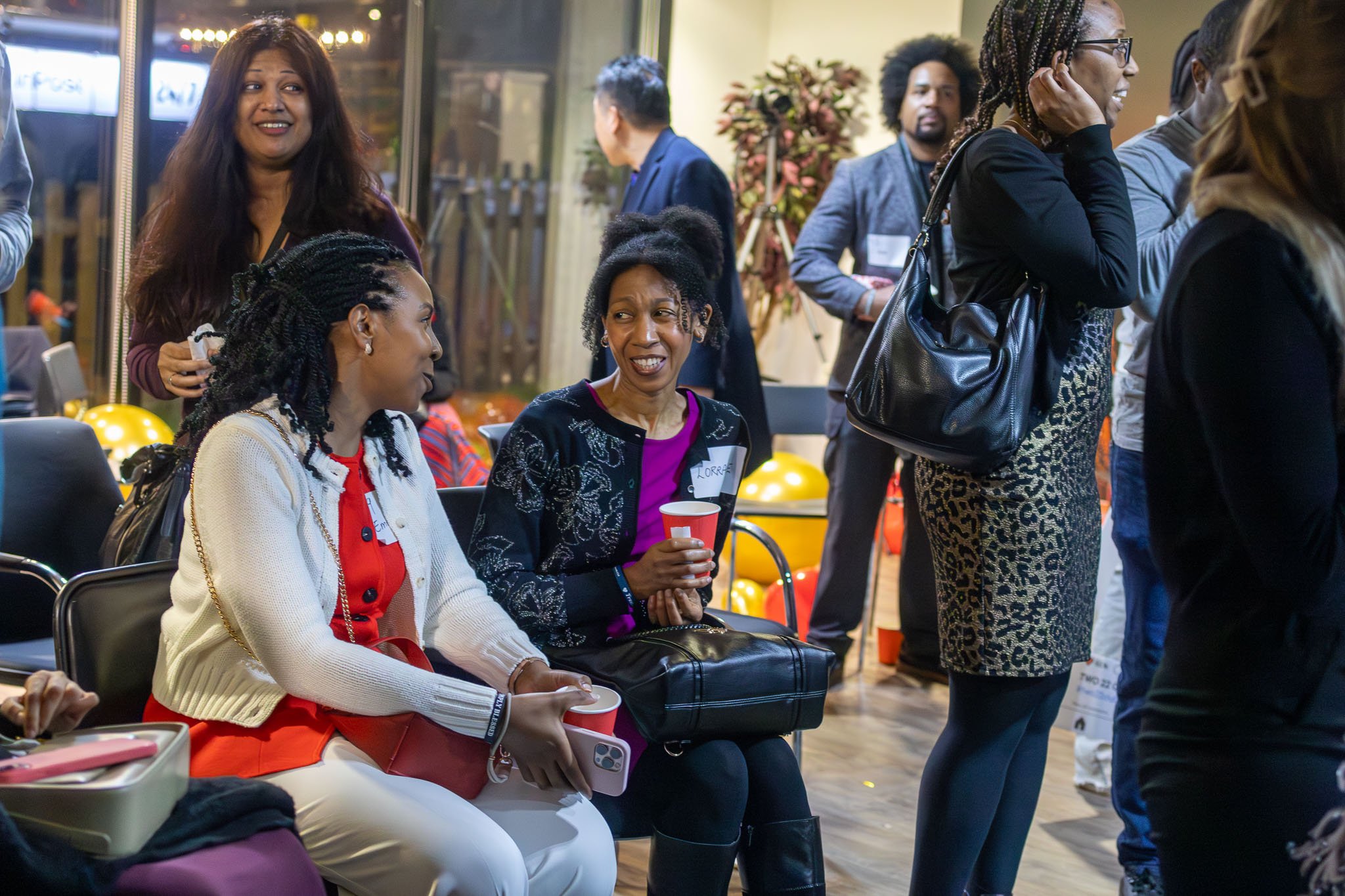 A group of diverse women at an event, some sitting and some standing, engaging in conversations and holding cups in an indoor setting with balloons and decorations.