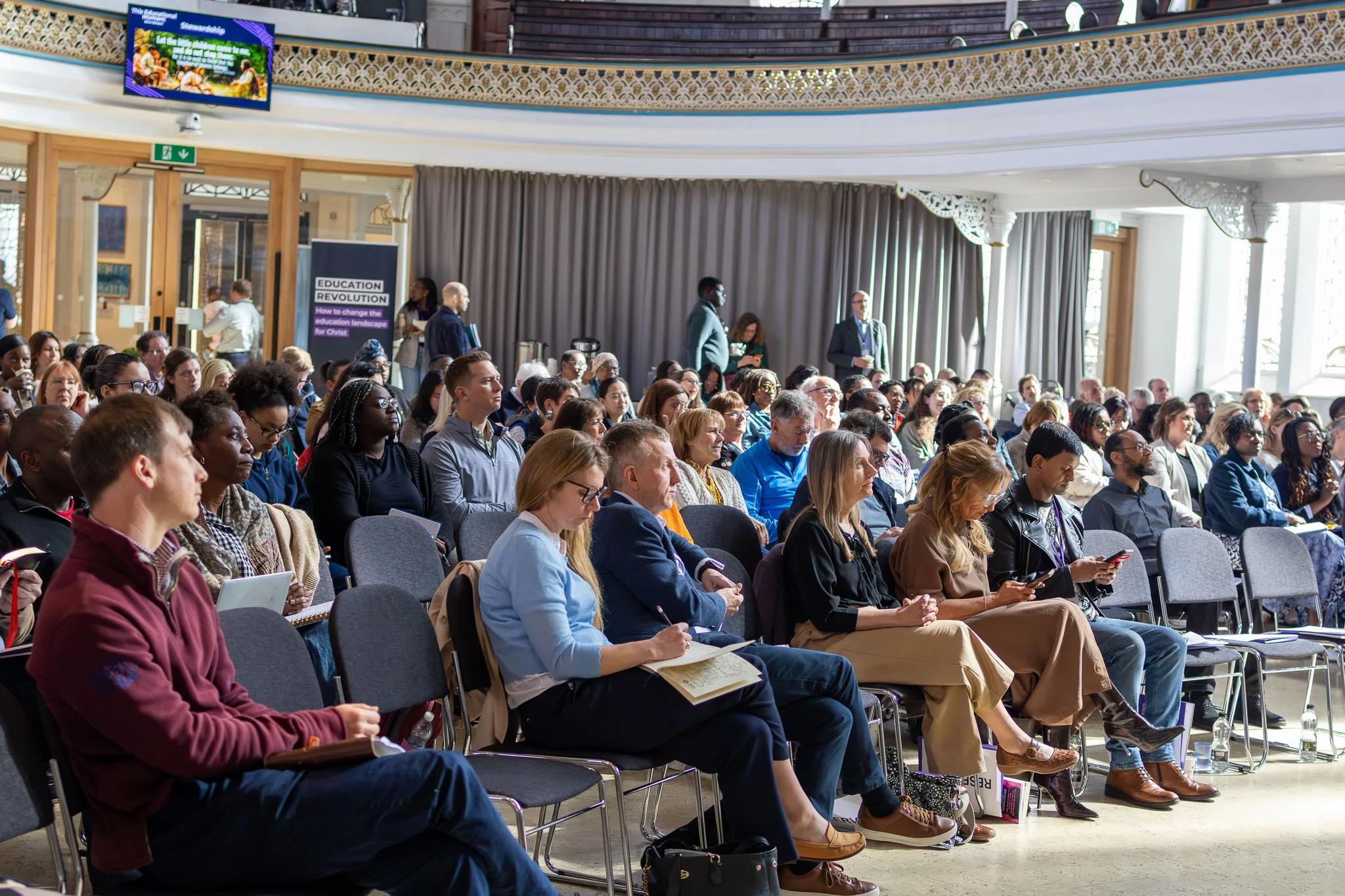 A large group of people attending a conference or seminar inside a building with ornate decor, large windows, and a stage area. Some are taking notes, others are listening attentively.