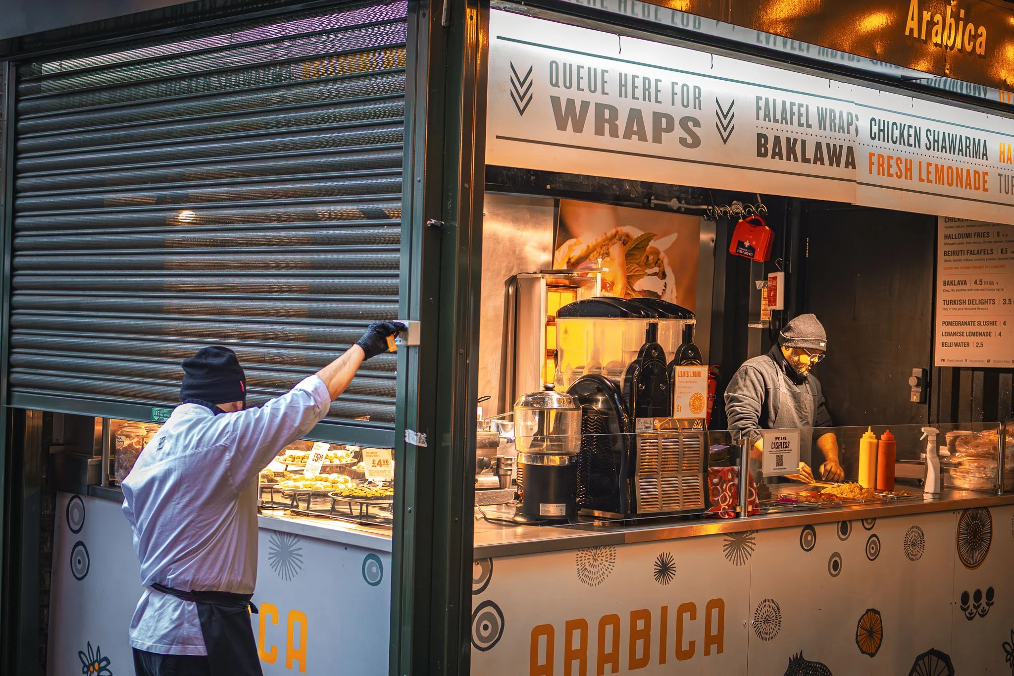 Food stand at Borough Market selling wraps, falafel wraps, chicken shawarma, baklava, and fresh lemonade. A man in a white shirt and black beanie is reaching inside the stand, while a woman in a black hoodie and glasses is preparing food inside.