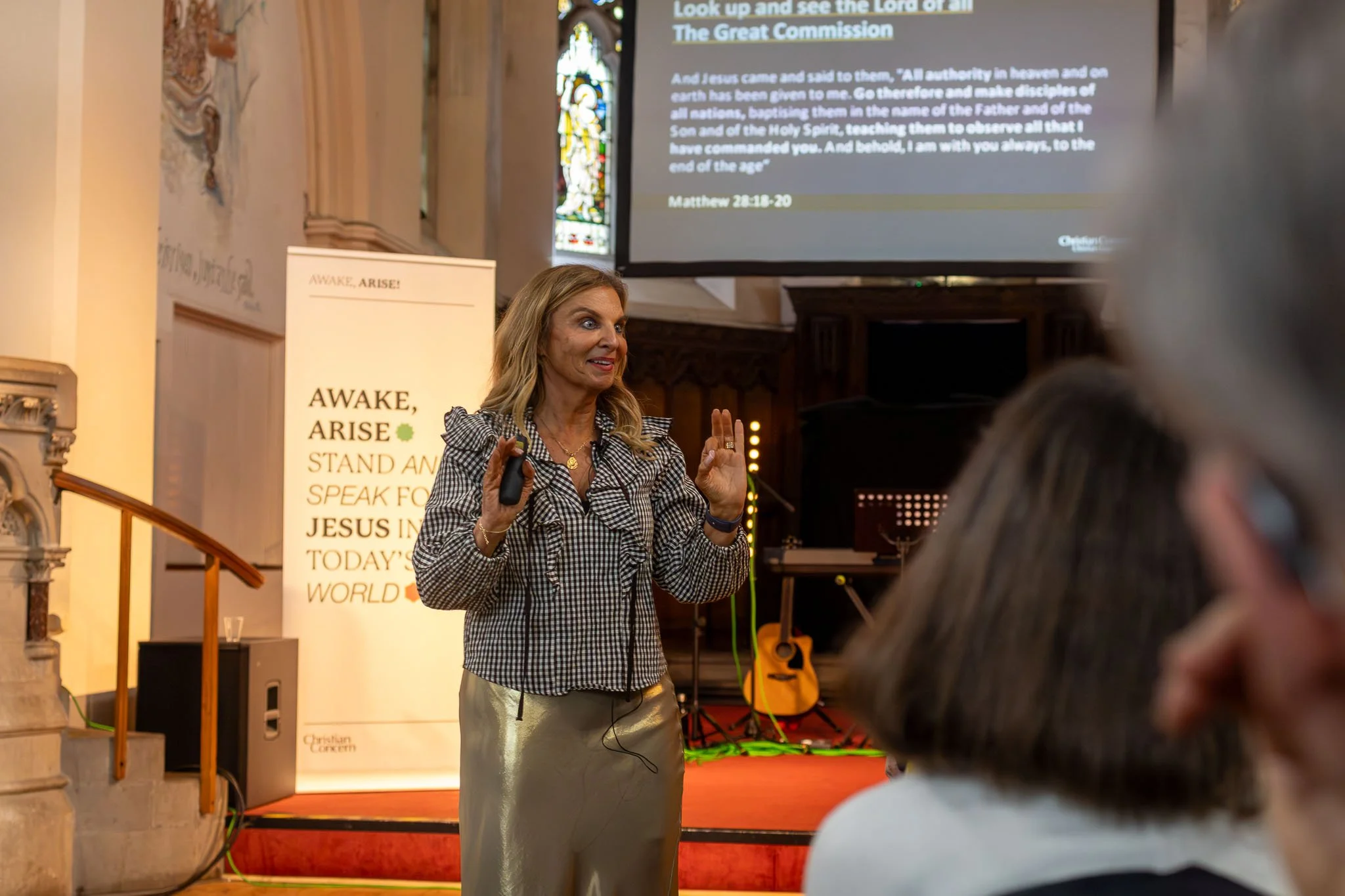 A woman in a checkered blouse and beige skirt speaking to a group in a church, with a large screen displaying a Bible verse in the background. Several people are seated in front of her, listening.