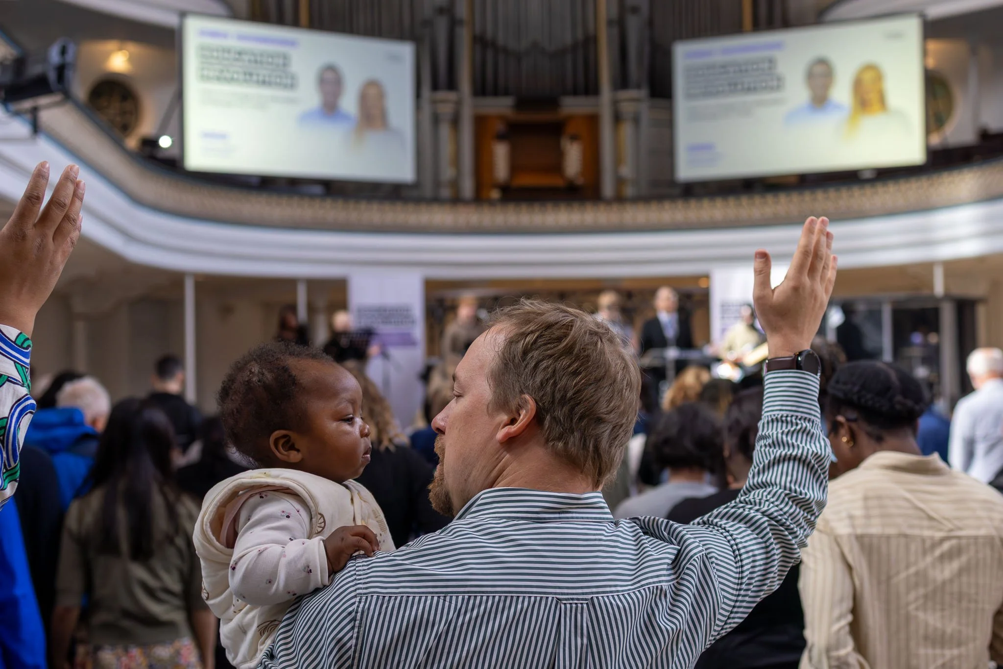 A crowd of people gathered indoors, with several raising their hands. In the background, two large screens display blurred images of a man and a woman. The setting appears to be a Christian event.