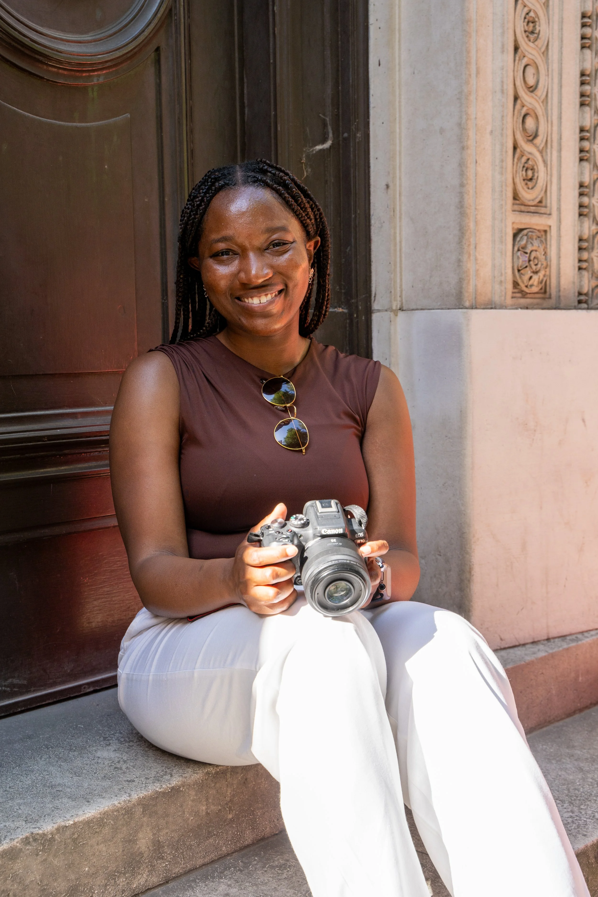 Rebeccah Onabajo with braided hair smiling and holding a camera, sitting on steps outside a building with ornate architecture.