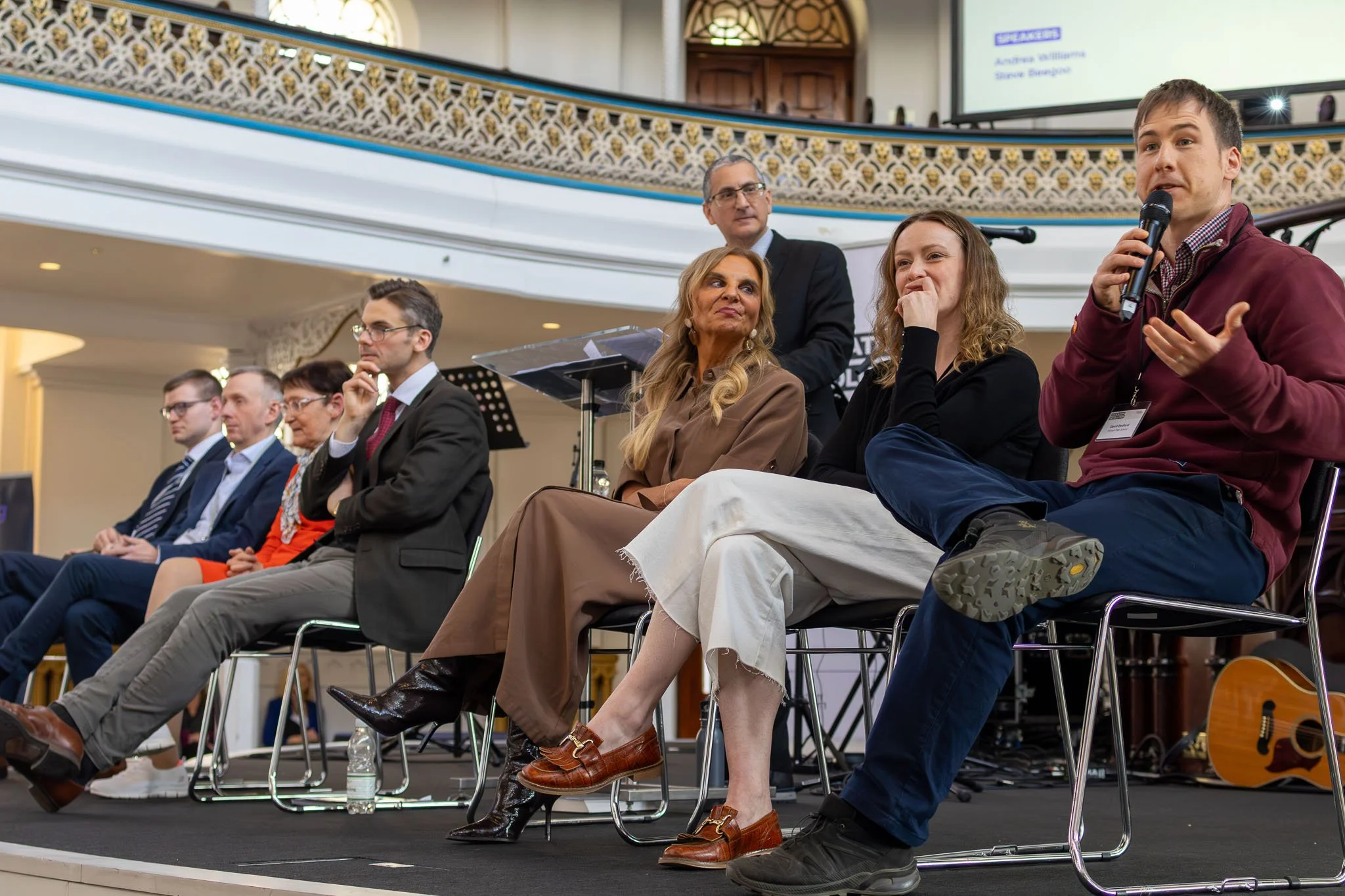A group of people seated on stage during a panel discussion or conference in an ornate indoor setting. One man is speaking into a microphone, while others listen attentively.