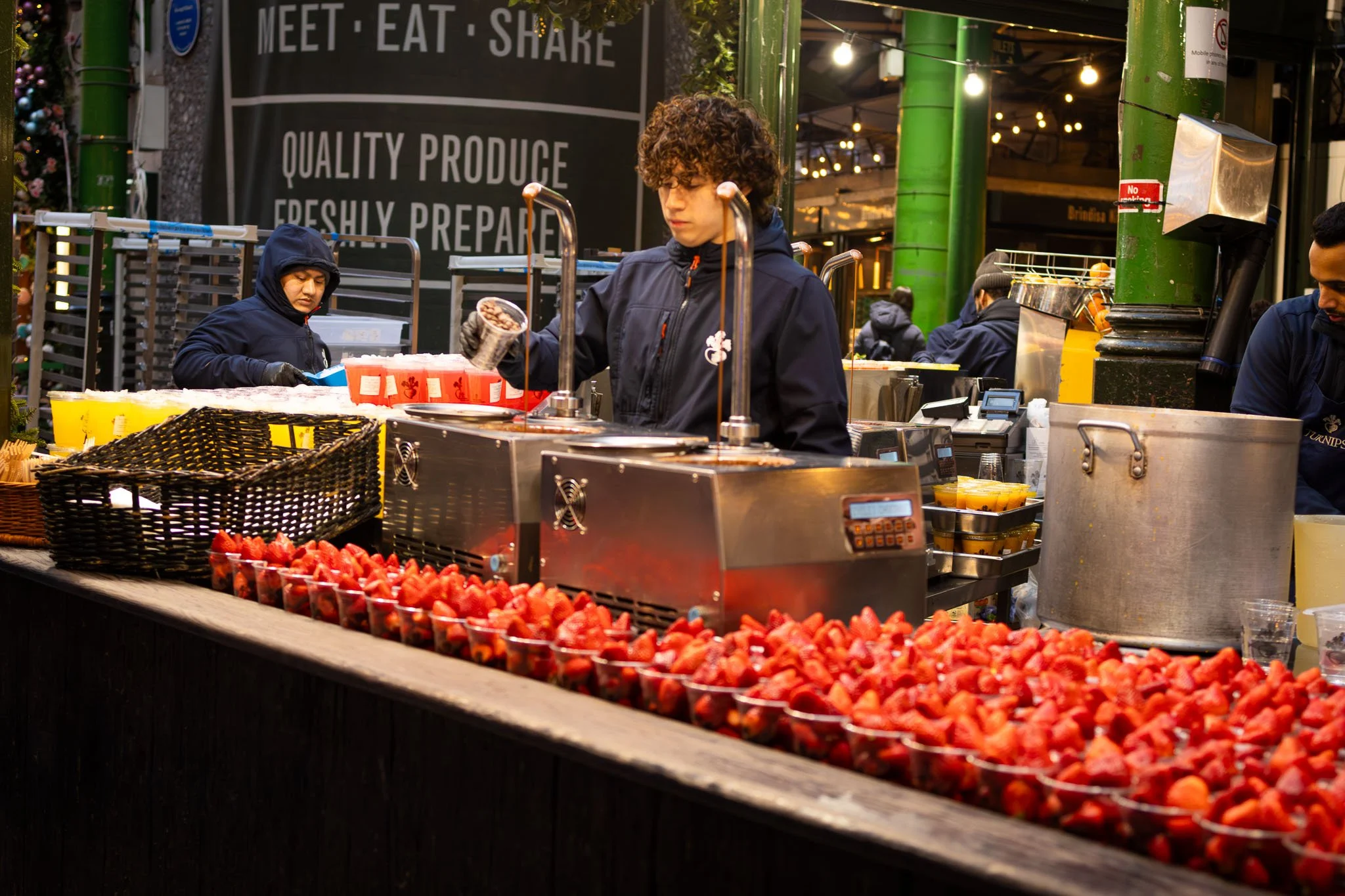 A street vendor stall at Borough Market with strawberries in the foreground and a person pouring a drink behind the counter. Sign in the background that reads 'Meet Eat Share' and other people visible in the background.