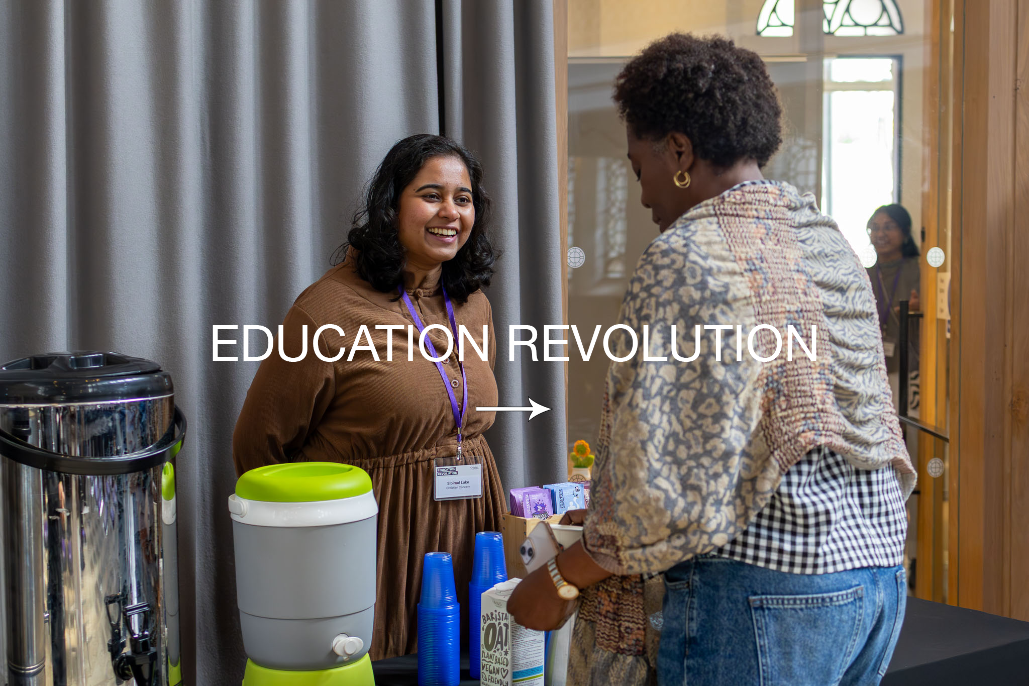 Two women having a conversation at a booth labeled 'Education Revolution' at an event. One woman, with short curly hair and gold hoop earrings, is facing away from the camera. The other woman, with medium-length dark hair, is smiling and wearing a brown dress with a lanyard. The booth has a water dispenser, blue cups, and informational materials. In the background, there is a woman with glasses smiling. The setting appears to be an indoor conference.