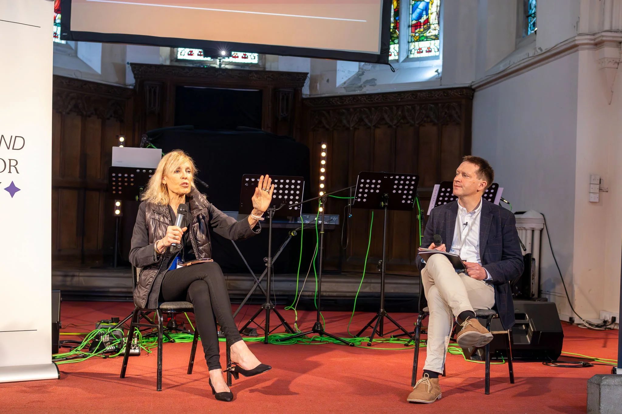 A woman with blonde hair, wearing a black leather jacket, sitting on a chair, holding a microphone, and gesturing with her hand. A man with short hair, wearing a dark blazer and beige pants, sits next to her, holding a notebook and a microphone. They