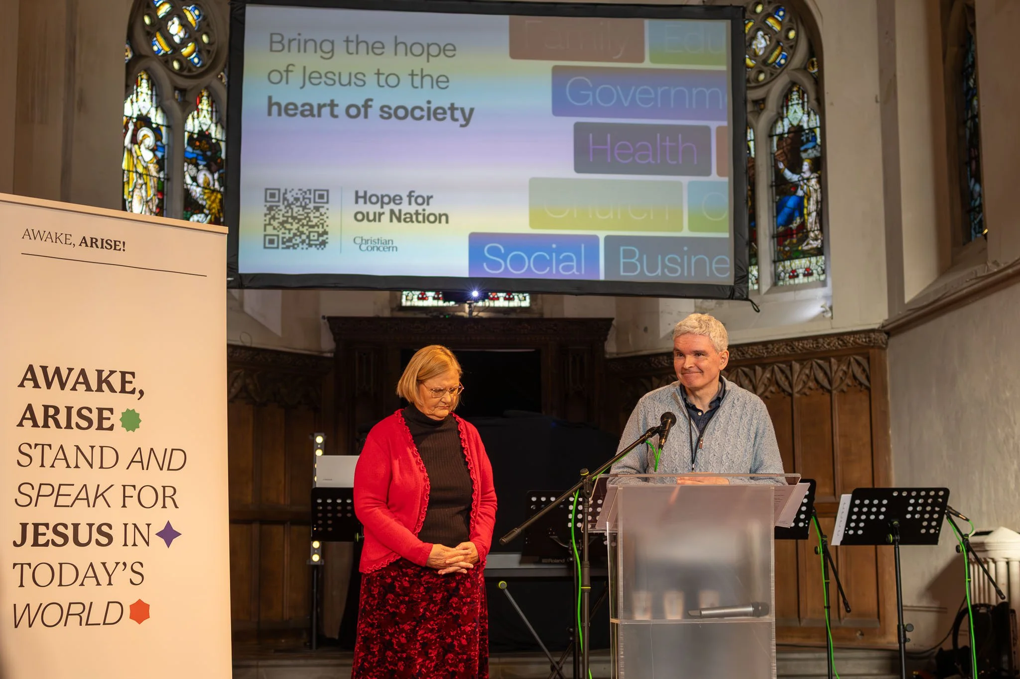 Women and man standing at a podium and microphone in a church with stained glass windows, with a large screen behind them displaying text about bringing hope of Jesus to society, and a sign with religious message on the left.