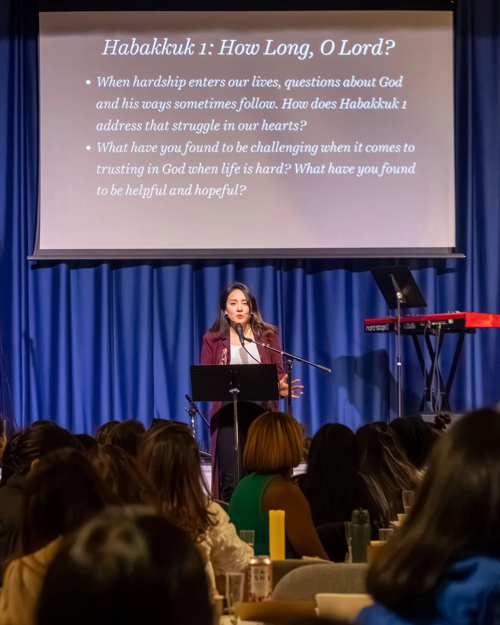 A woman is giving a speech or presentation at a stage with a microphone. Behind her, there is a projection screen displaying a slide titled 'Habakkuk 1: How Long, O Lord?' with two bullet points. The first bullet asks about how hardship and questions