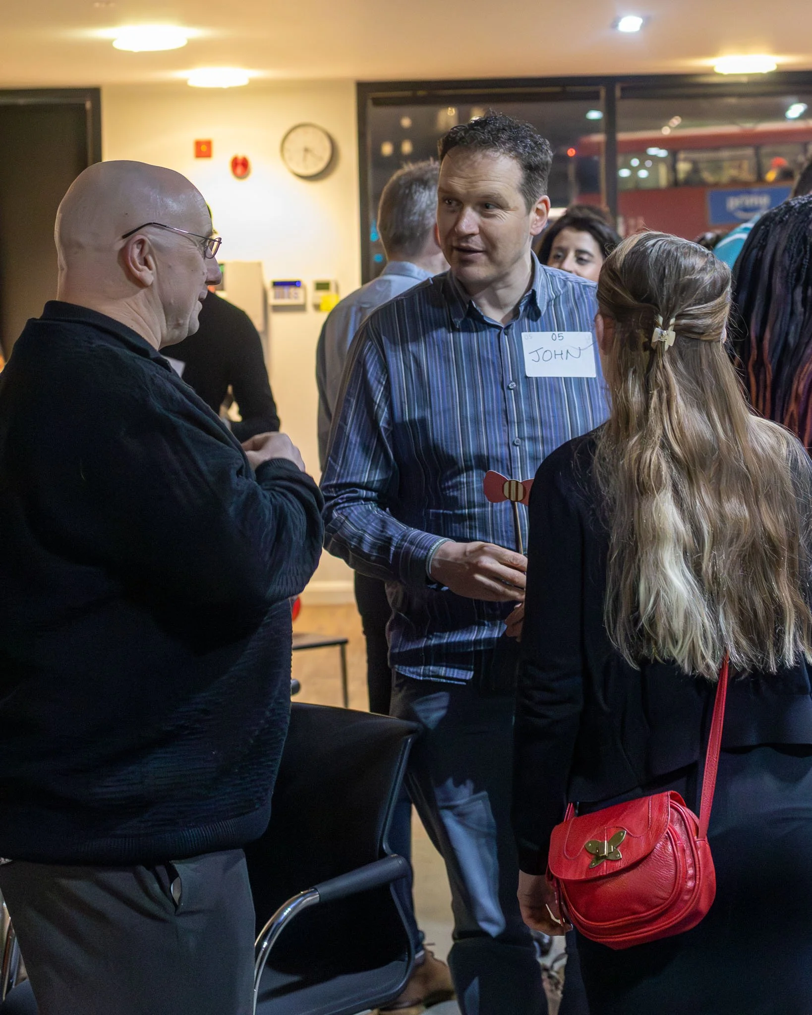 People socializing indoors, including a man with a name tag labeled 'John,' a woman with long blonde hair and a red purse, and an older man with glasses. The background features a clock and other people, with a visible bus outside the window.