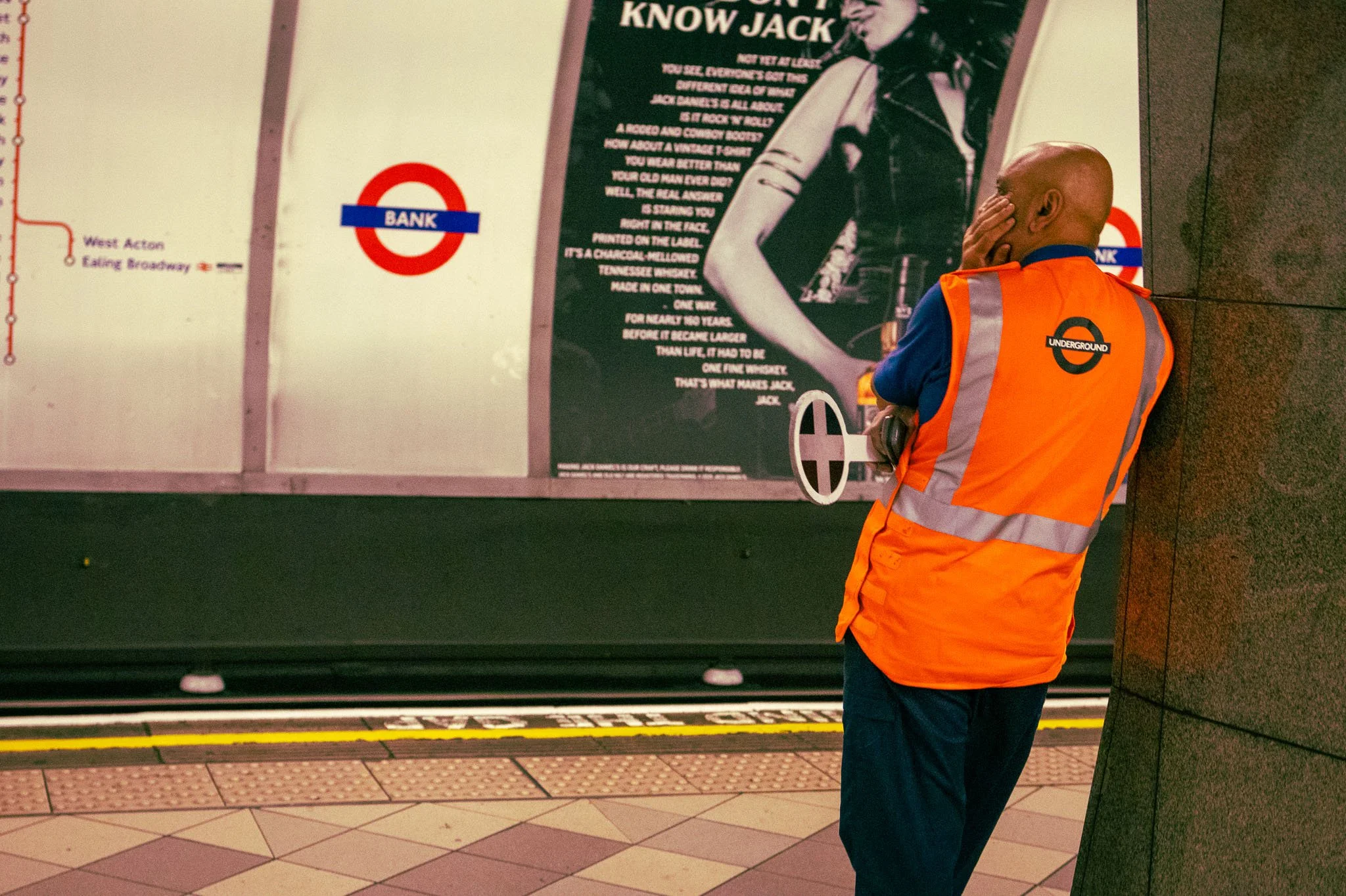 A man with no hair wearing a bright orange UK Underground safety vest is standing in Bank Station, facing a large advertisement poster. The man is covering his part of his face with his hand.