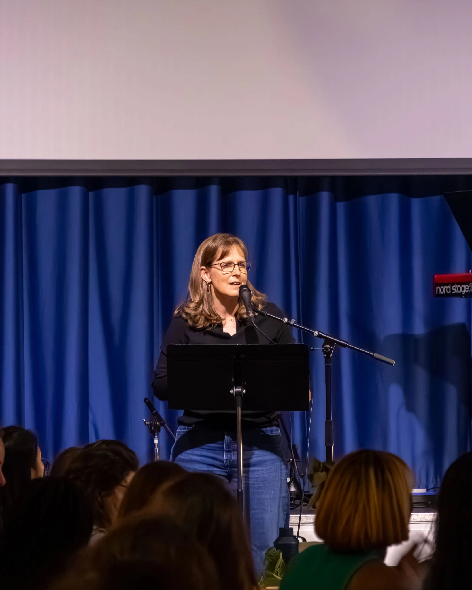 Woman with glasses speaking at a podium in front of a blue curtain during a presentation or event, with an audience in the foreground.