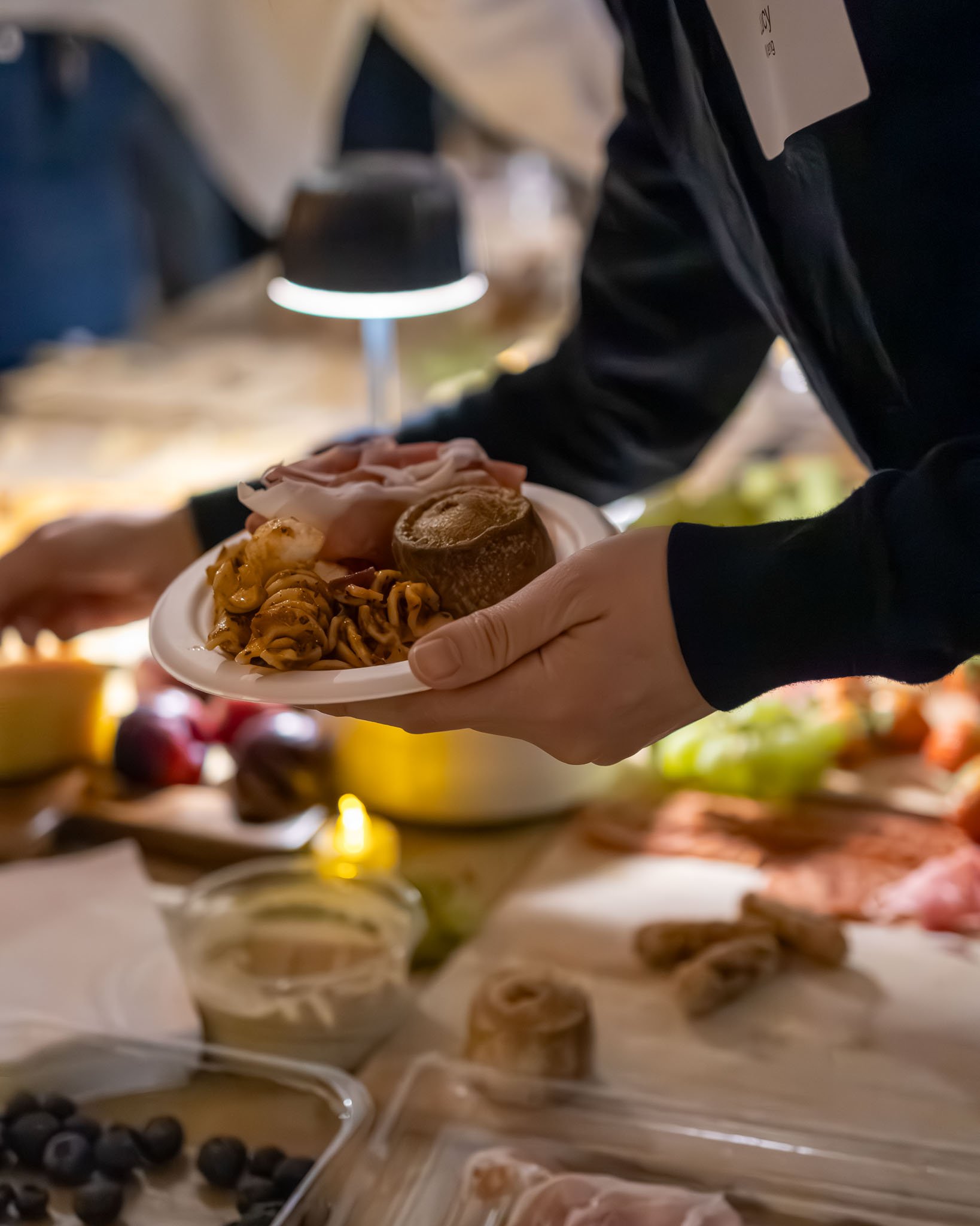 A person serving food on a plate with pasta, a roll, and slices of meat or cheese, at a church with various dishes in the background.