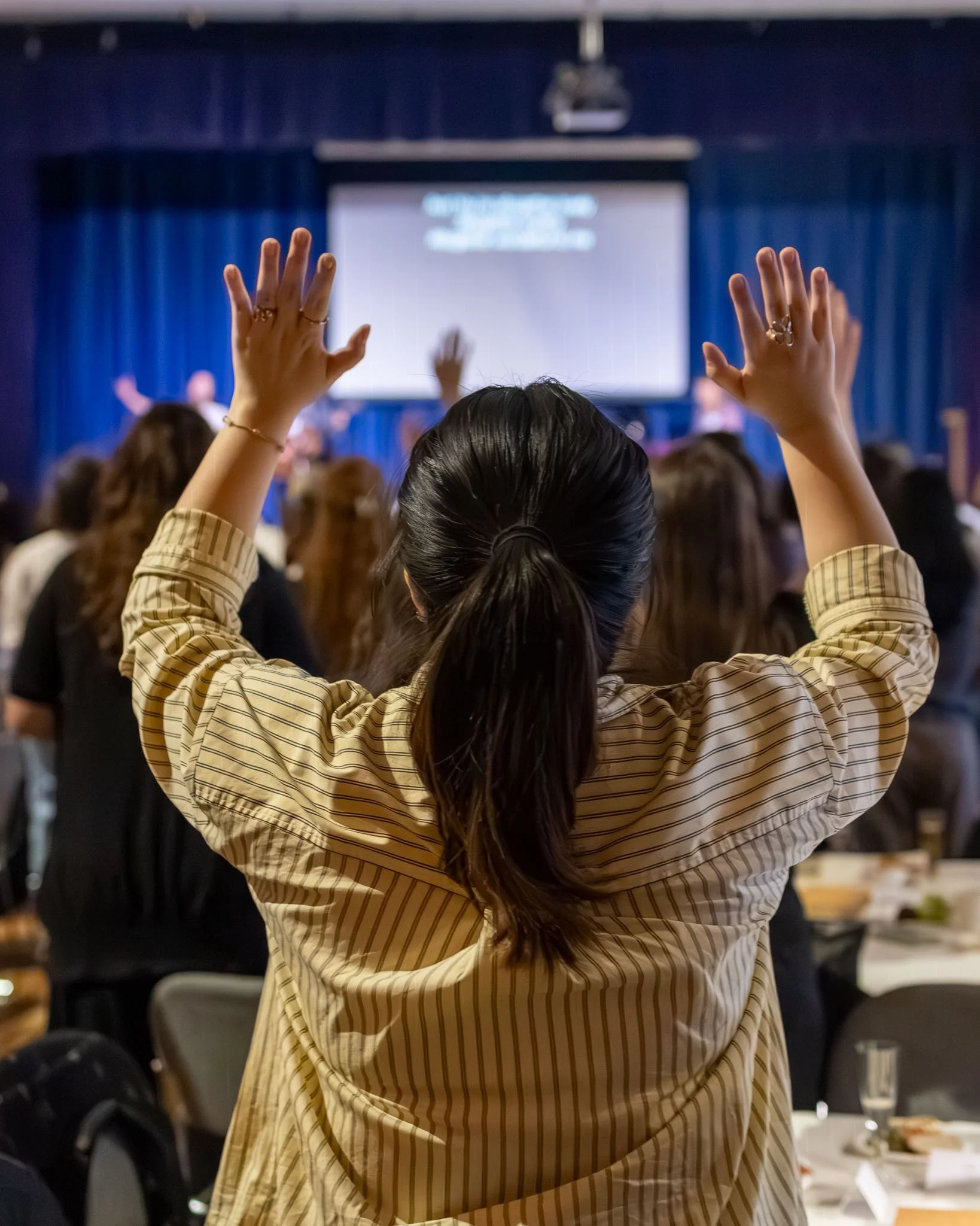 A woman with dark hair tied back, wearing a beige and brown striped shirt, raises her hands with fingers apart while attending an event in a room with blue curtains, a projector screen at the front, and other attendees.