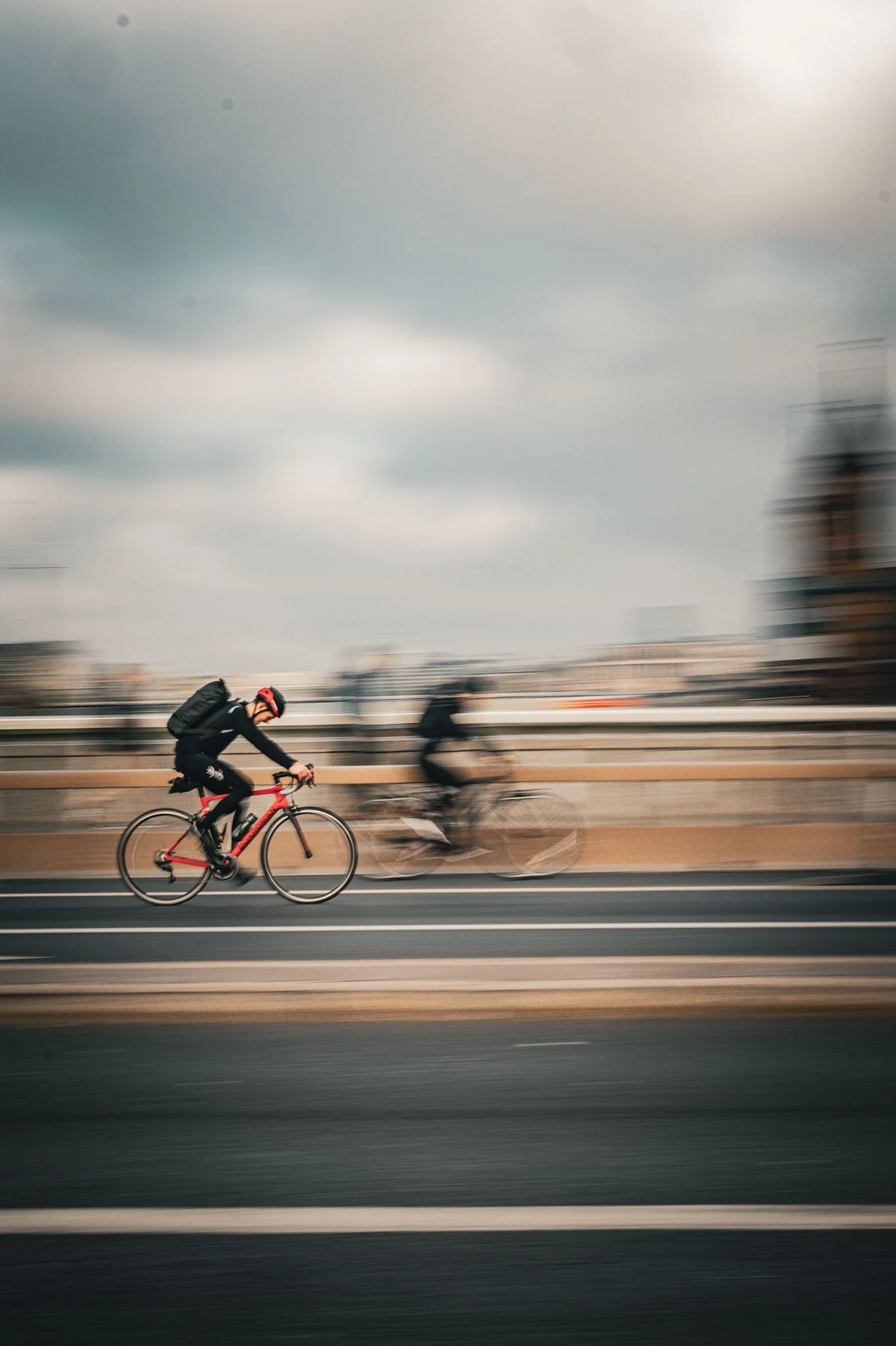 Blurred motion of a cyclist wearing a black outfit and helmet, riding a red bicycle on a city street with cloudy sky and tall buildings in the background.