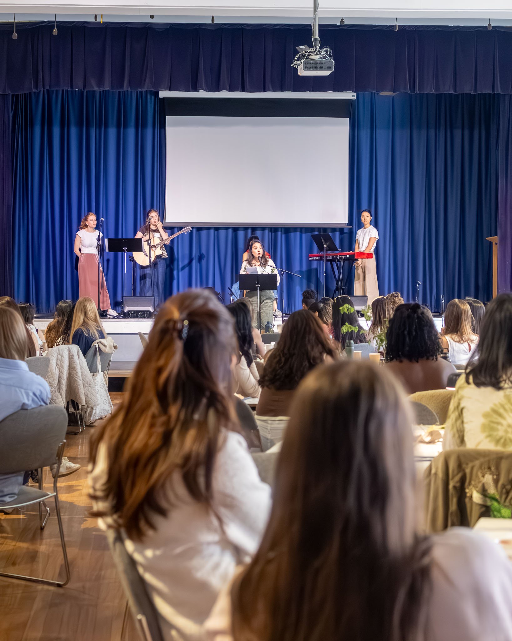 A group of young women performing on a stage in front of an audience. One is playing guitar, another singing, and a third with a keyboard. The stage is decorated with blue curtains and a large blank white screen.