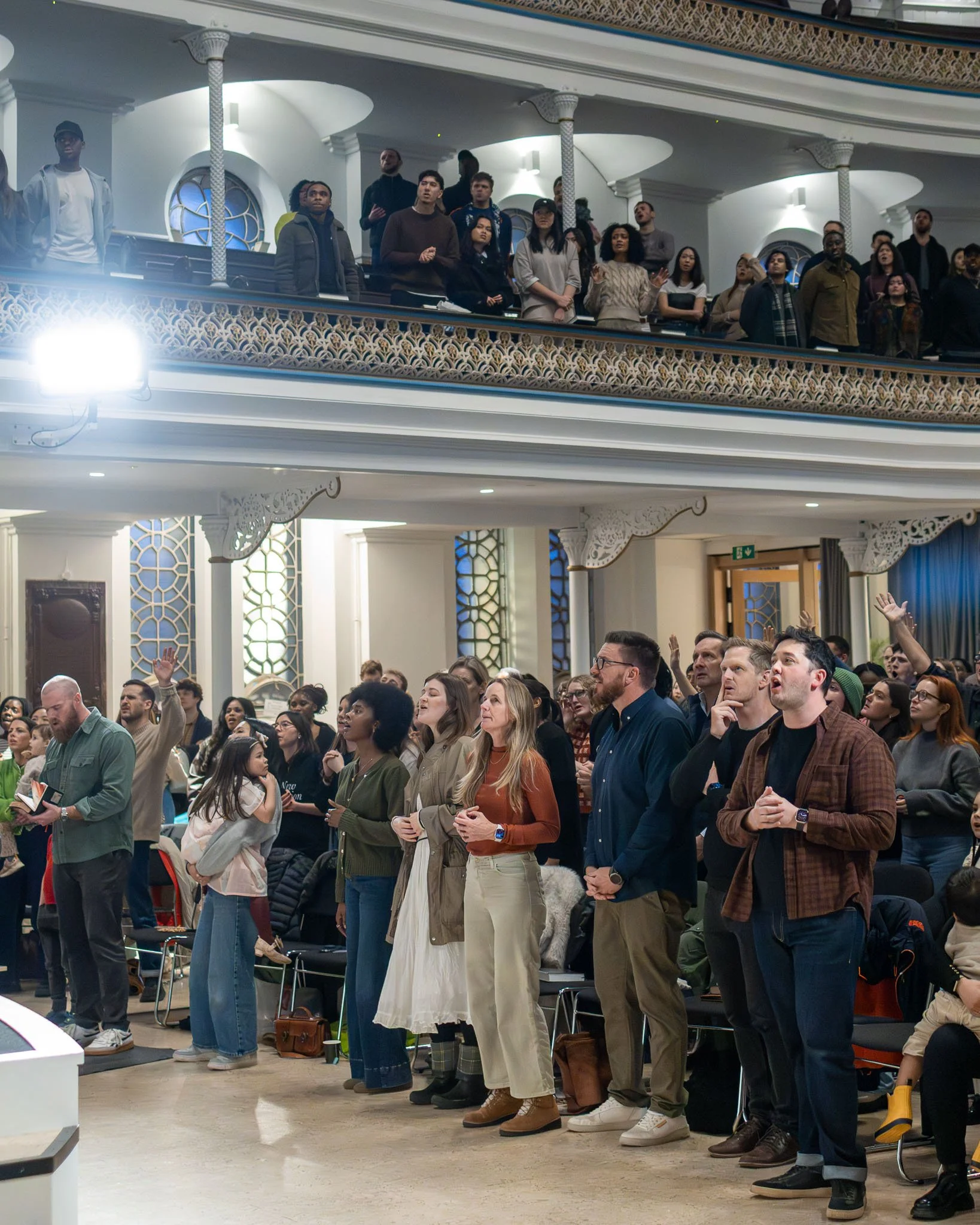 A large group of diverse people standing and praying inside a church or auditorium during a religious gathering. They are facing forward with some having their eyes closed and hands raised.