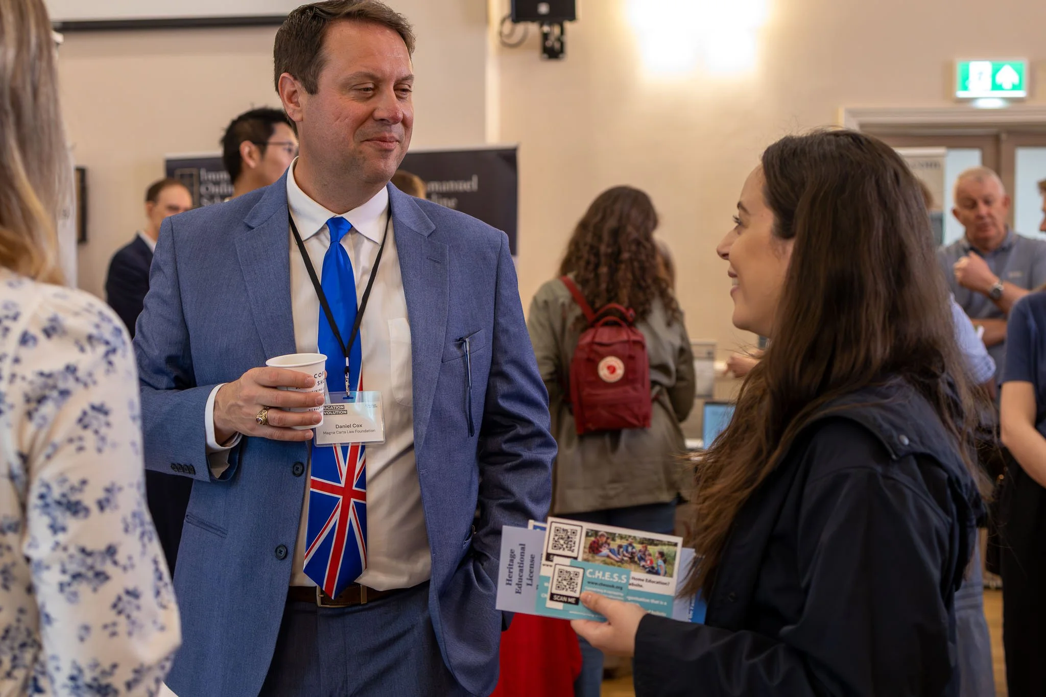 A professional man in a blue suit and Union Jack tie holding a white cup, engaging in conversation with a woman at a conference or event. The woman is holding informational brochures. There are other people in the background, and the setting appears 
