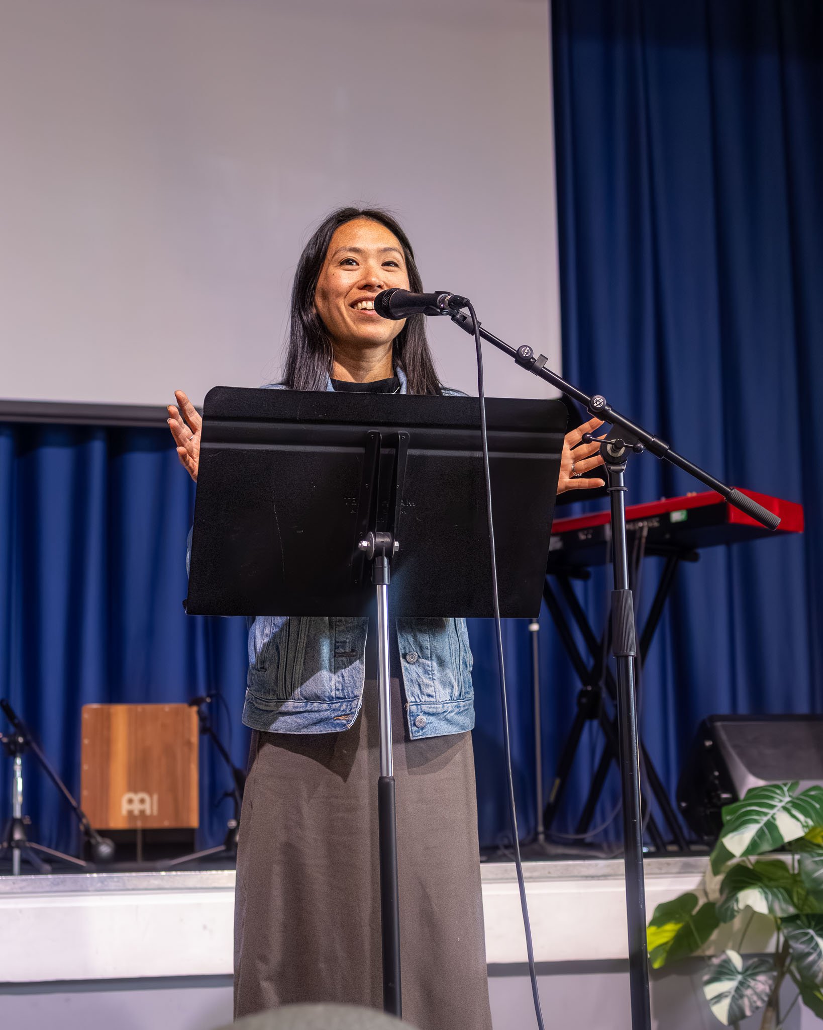 A woman standing at a podium with a microphone, smiling, wearing a denim jacket and a long skirt, on a stage with blue curtains and musical instruments in the background.