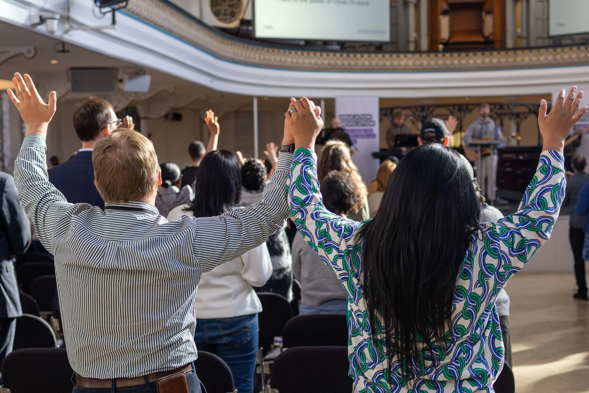 People attending a conference, raising their hands during a session, in a large, ornate indoor venue.
