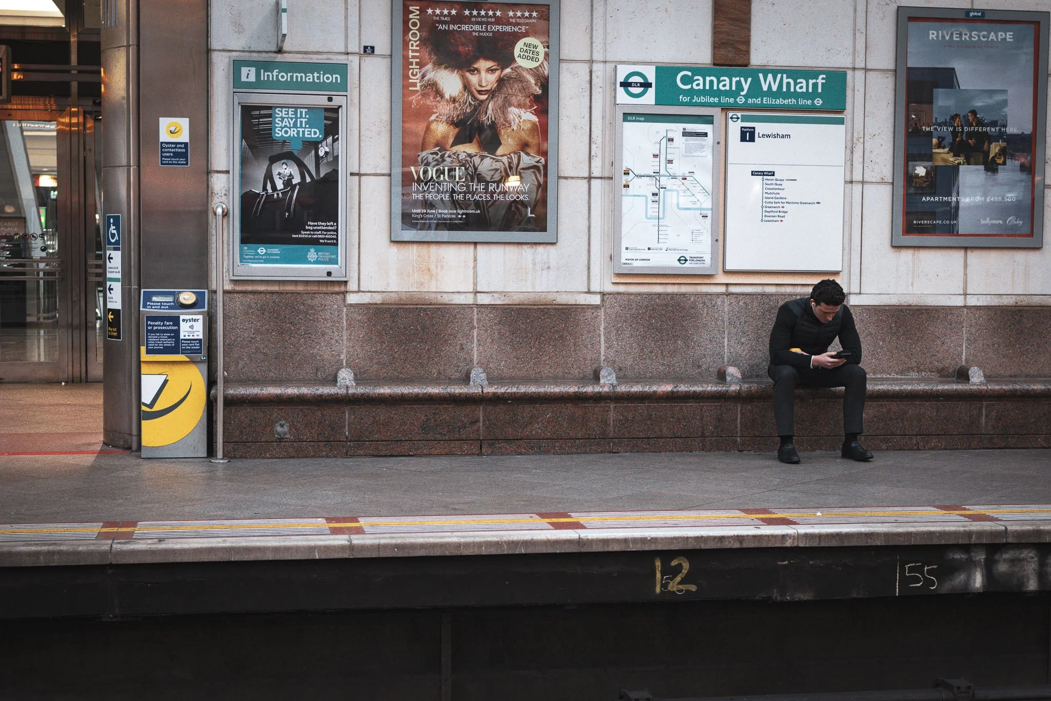 A man in a black coat and black shoes sitting on a bench at Canary Wharf DLR station, looking at his phone. Behind him is a wall with signs and posters, including transportation maps, advertisements, and information boards.
