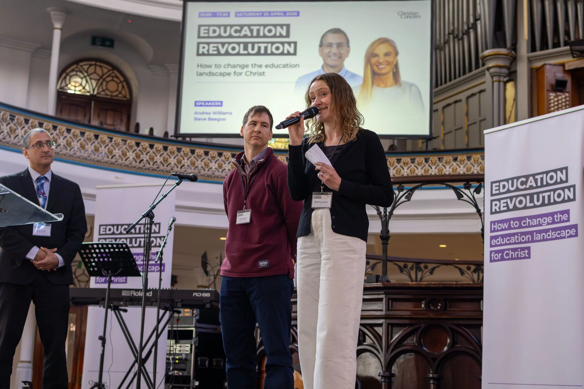 At a conference, a woman is speaking into a microphone while standing in front of a large screen that displays the title "Education Revolution" and mentions a discussion on changing the education landscape for Christ. There are a few men standing nea