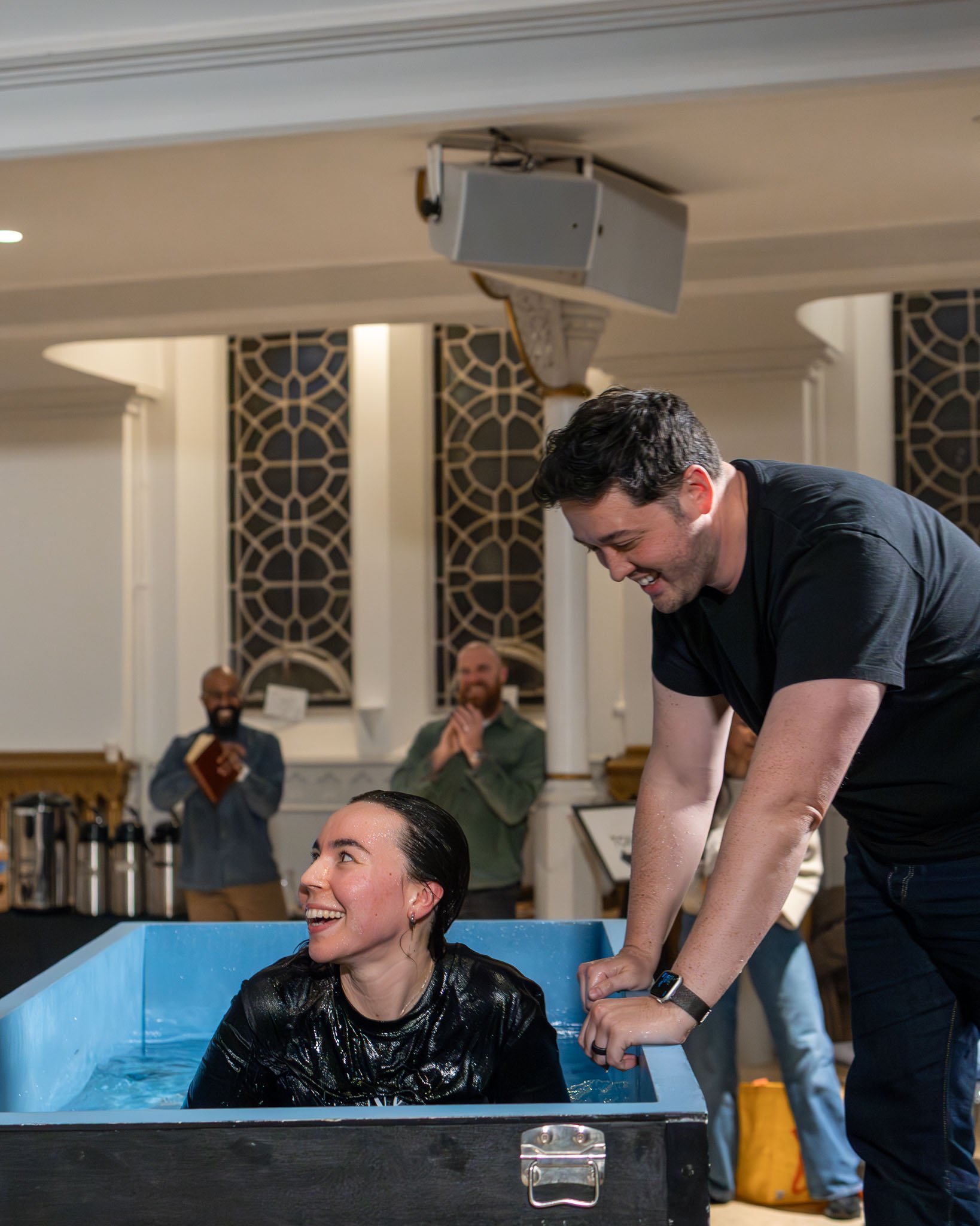 A woman getting baptized in a church or religious setting while a man holds her hands and supports her in a baptismal pool. Two men, one holding a book, are seen clapping and smiling in the background.