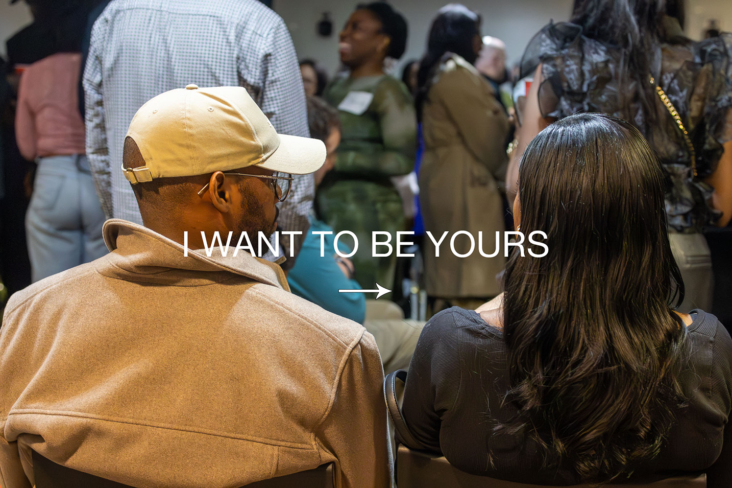 Two people sitting and talking at a crowded event, with the text 'I WANT TO BE YOURS' overlaid.