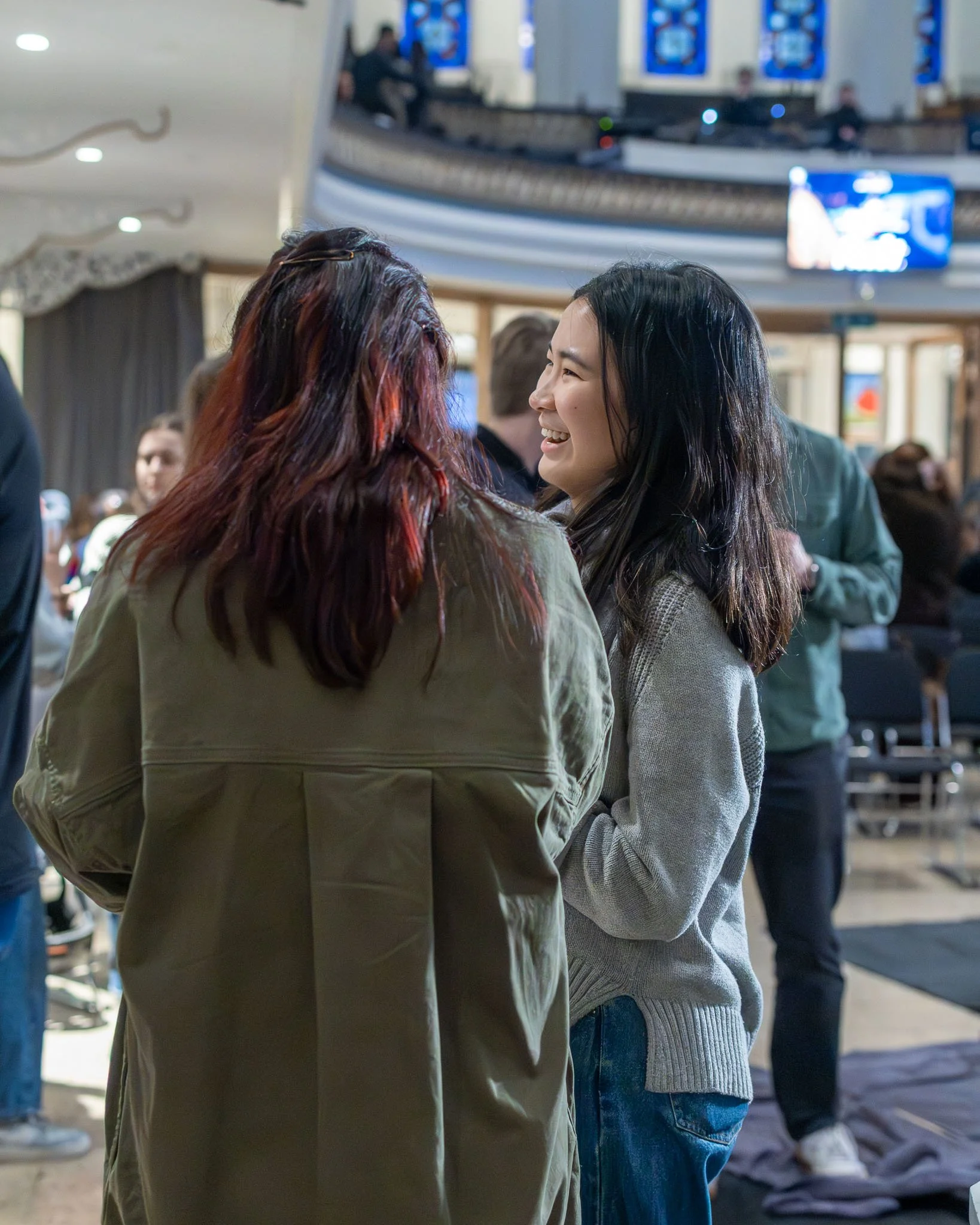 Two women engaged in conversation at a busy indoor venue with multiple people and screens in the background.