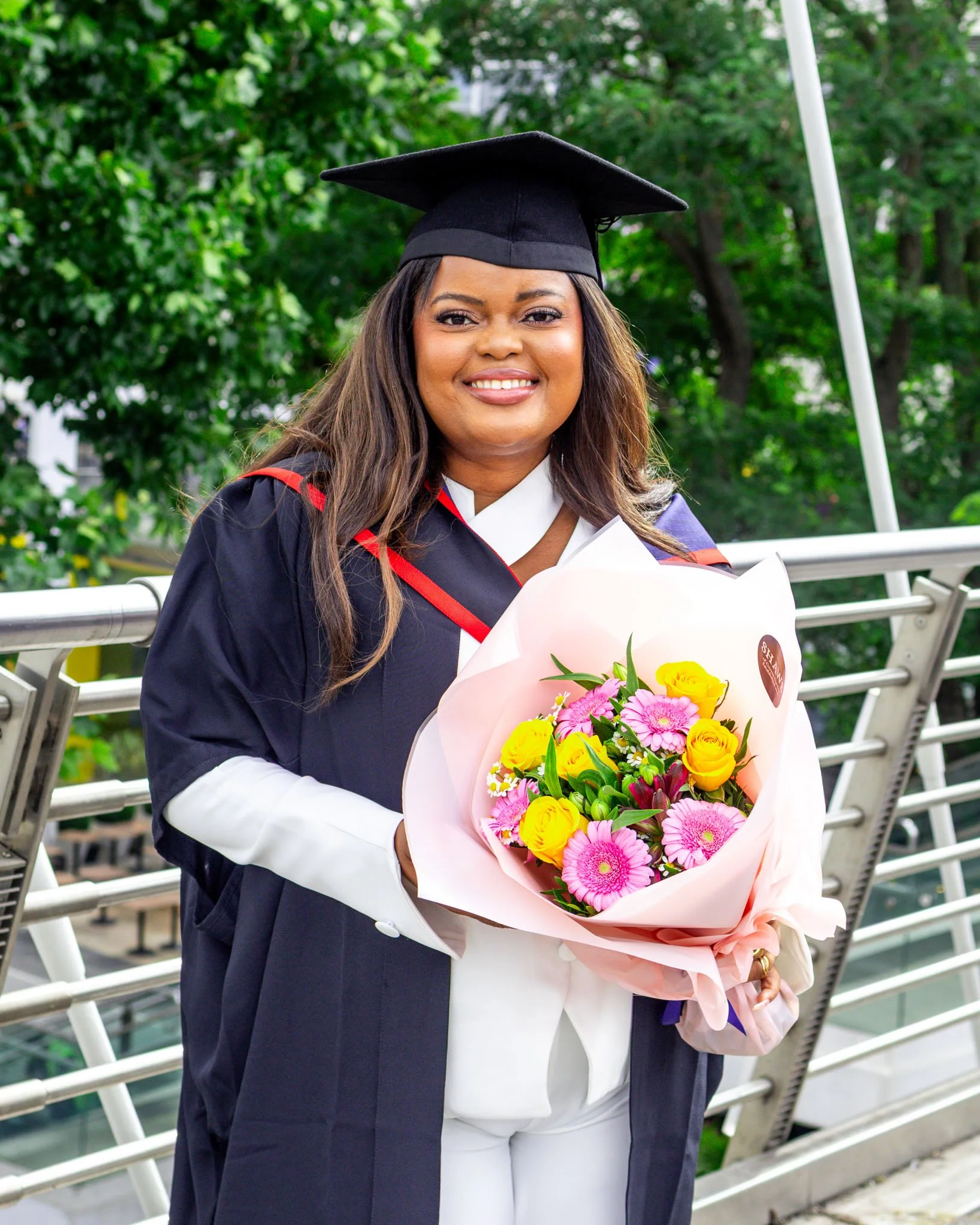 A woman wearing a graduation cap and gown holding a colorful bouquet of flowers outdoors, with green trees in the background.