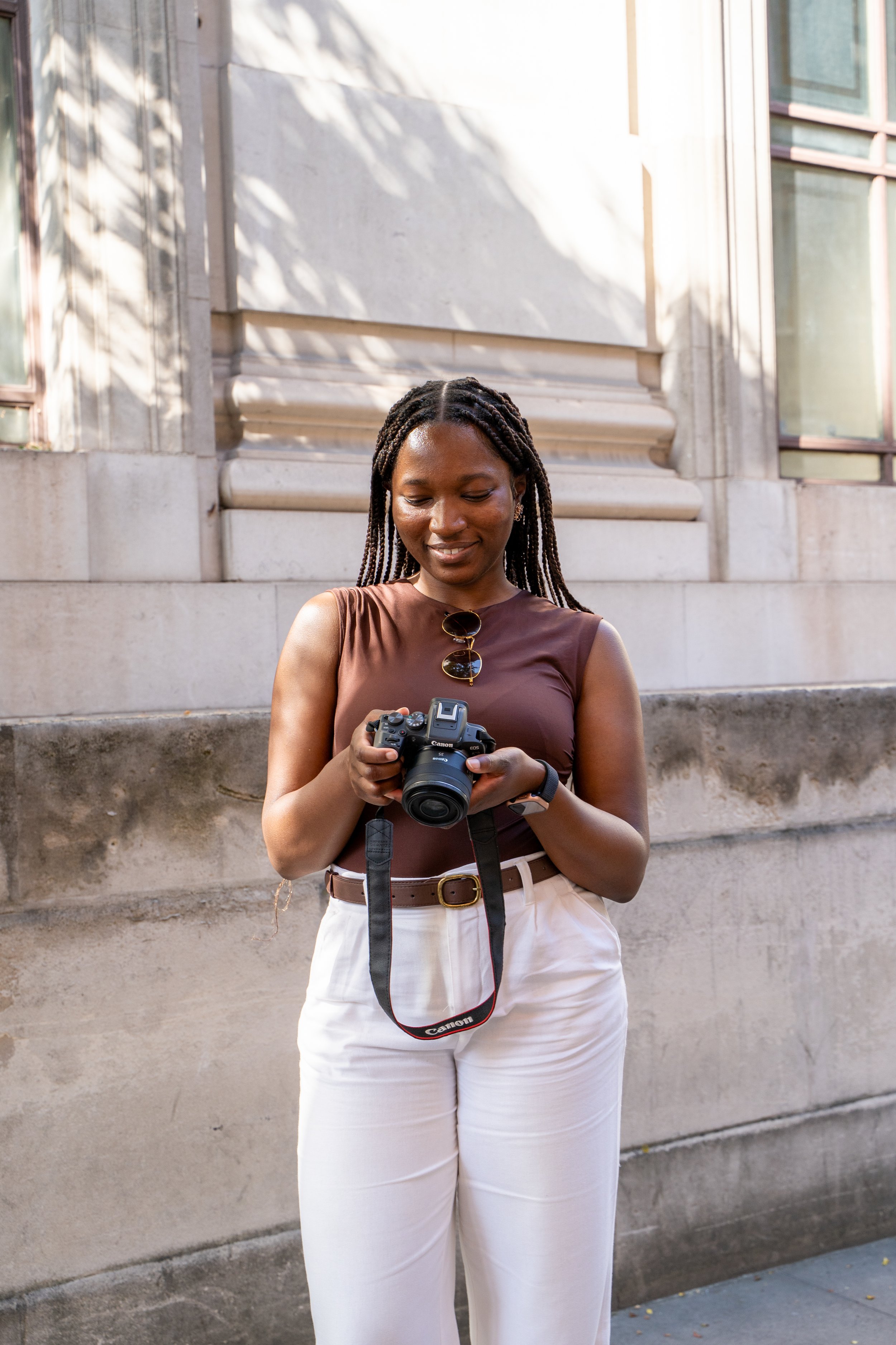 Rebeccah Onabajo standing in front of a stone building, looking down at a camera she is holding in her hands. She is wearing a sleeveless brown top, white pants, and has sunglasses hanging from her top. She has braided hair and is smiling.