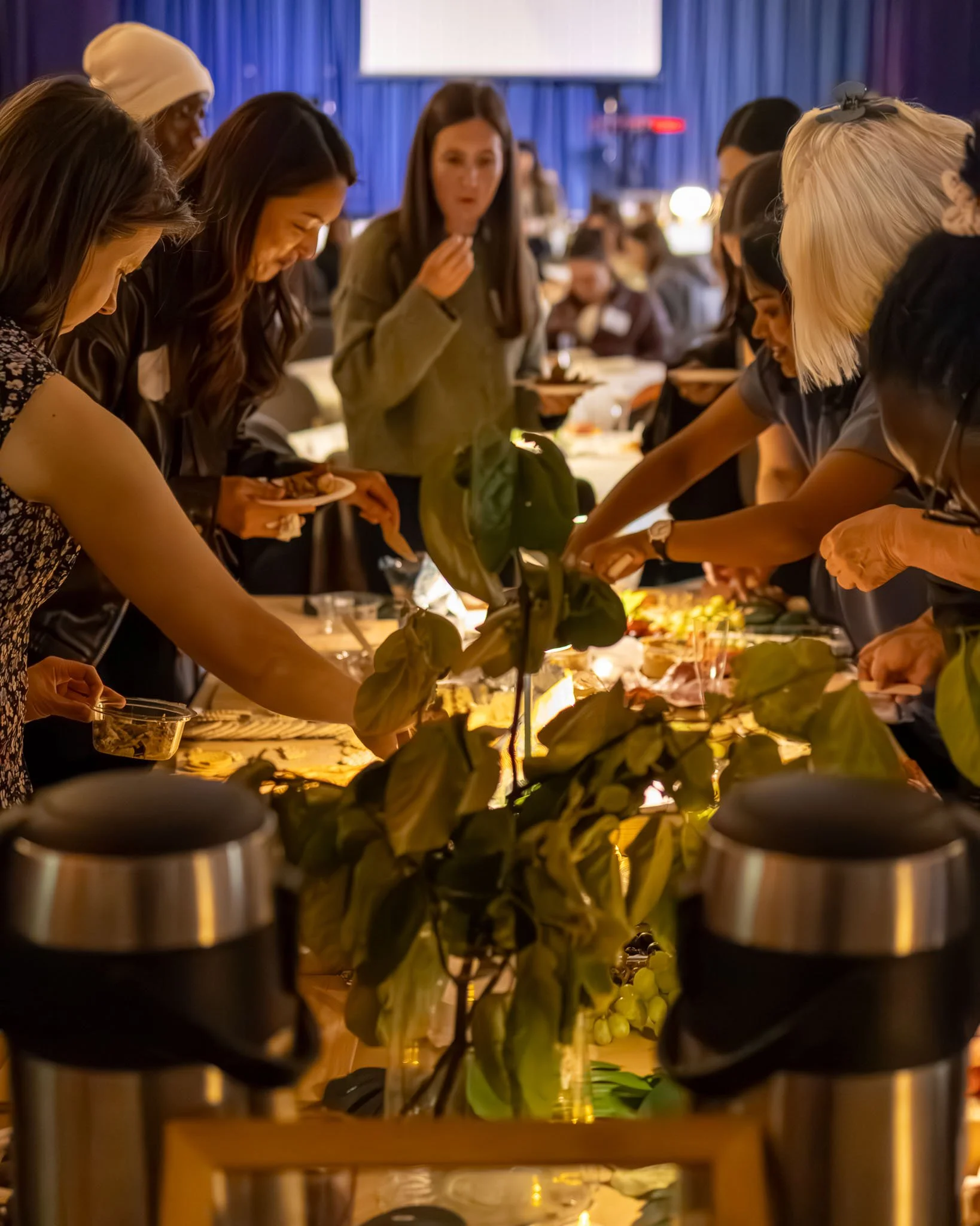 People gathered around a buffet table, serving themselves food. The scene appears to be at a church with a blue curtain backdrop.