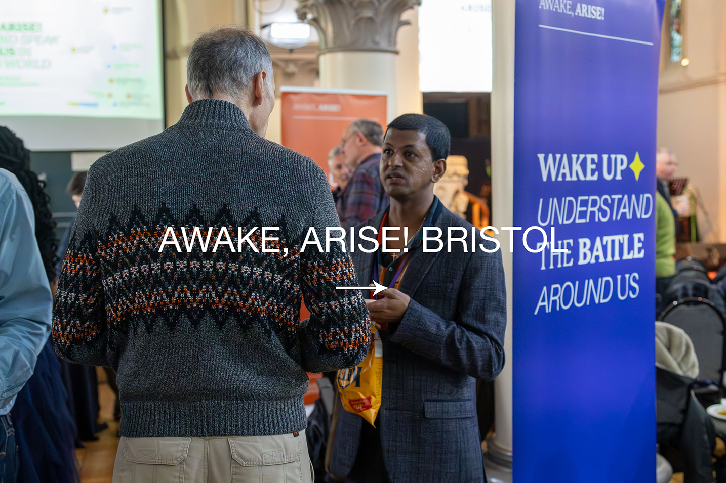 Two men are engaged in conversation at an event, one seen from the back wearing a dark sweater with a patterned design, and the other facing forward in a dark blazer, with a lanyard around his neck. A large blue banner in the background reads 'WAKE UP UNDERSTAND THE BATTLE AROUND US'.