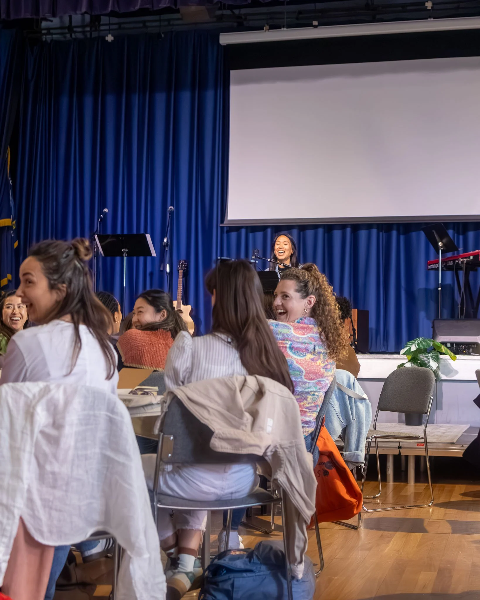 A group of young women seated at tables and smiling in a room with blue curtains, attending a church event, with a smiling woman on stage in front of a large white screen.