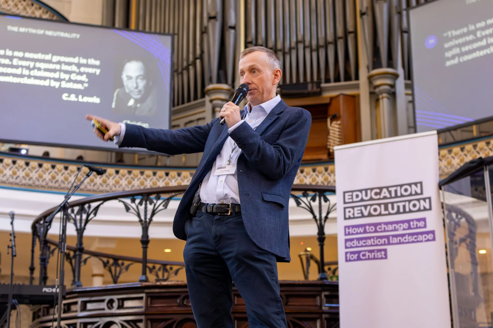 A man in a suit jacket, white dress shirt, and wearing a lanyard, giving a presentation with a microphone in front of a large screen displaying a quote about neutrality by C.S. Lewis, in a venue with ornate architectural details.