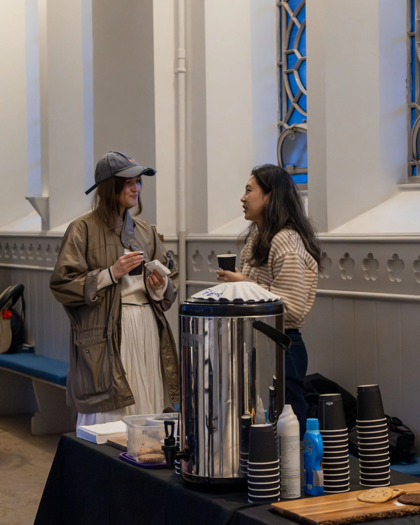 Two women standing and talking at a table with a drink in a cafe or event space. One woman wears a gray cap, tan jacket, and cream skirt. The other woman has long dark hair and wears a striped top. The table has cups, bottles, a cooler, and paper pro