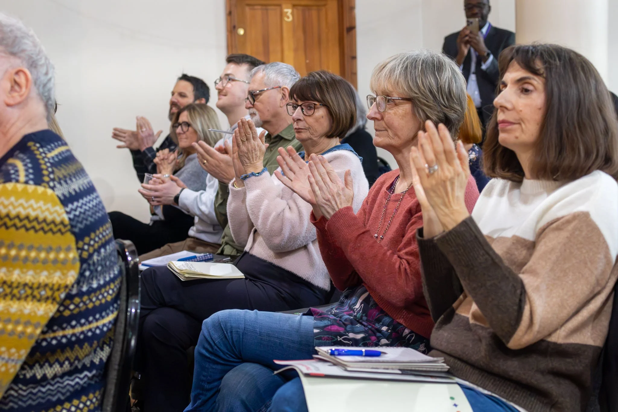 A group of diverse adults sitting in rows, attentively listening at a meeting or conference, with some taking notes.