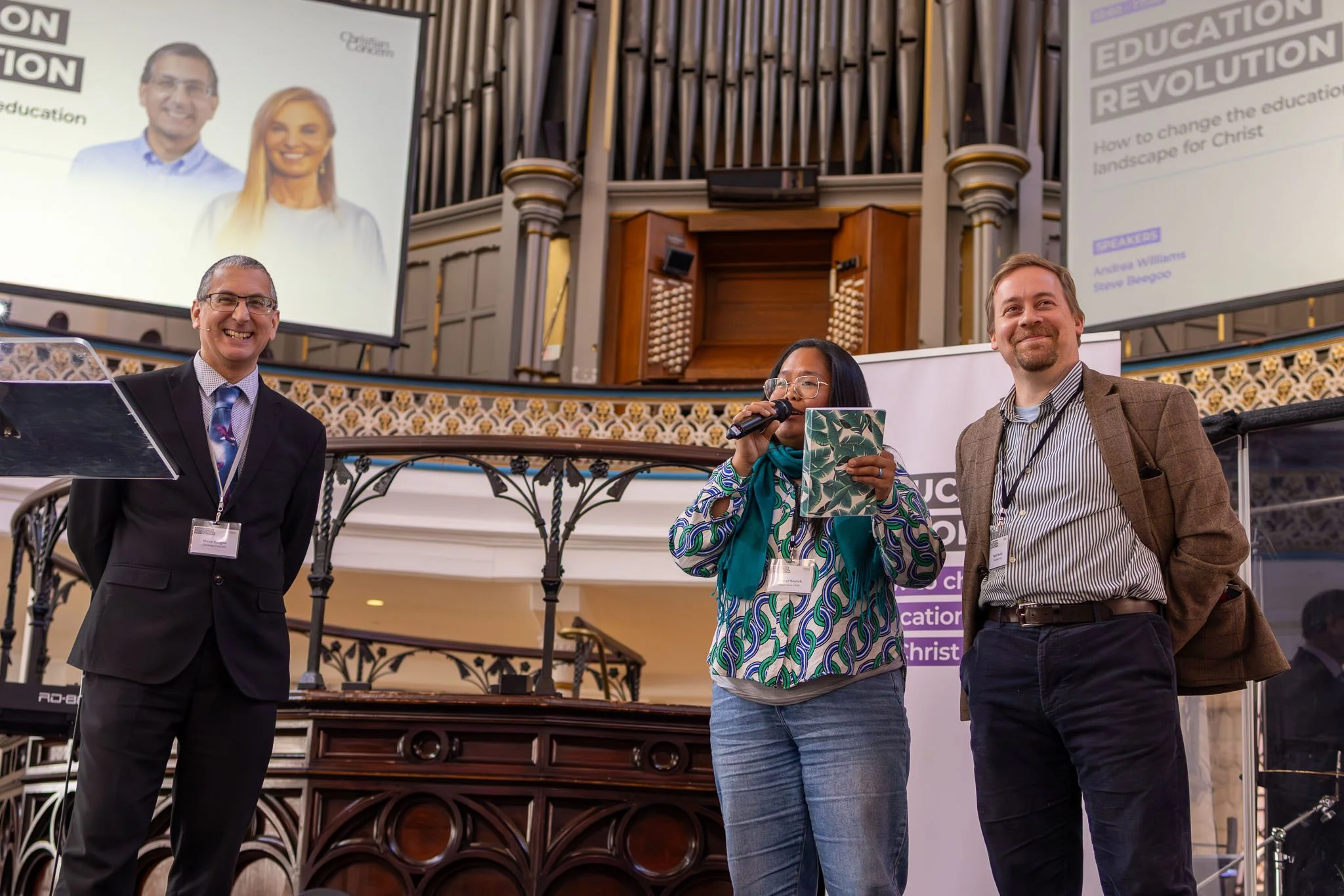Three people standing on a stage in front of a large screen and organ pipes, with banners about Christian education and a woman speaking into a microphone.