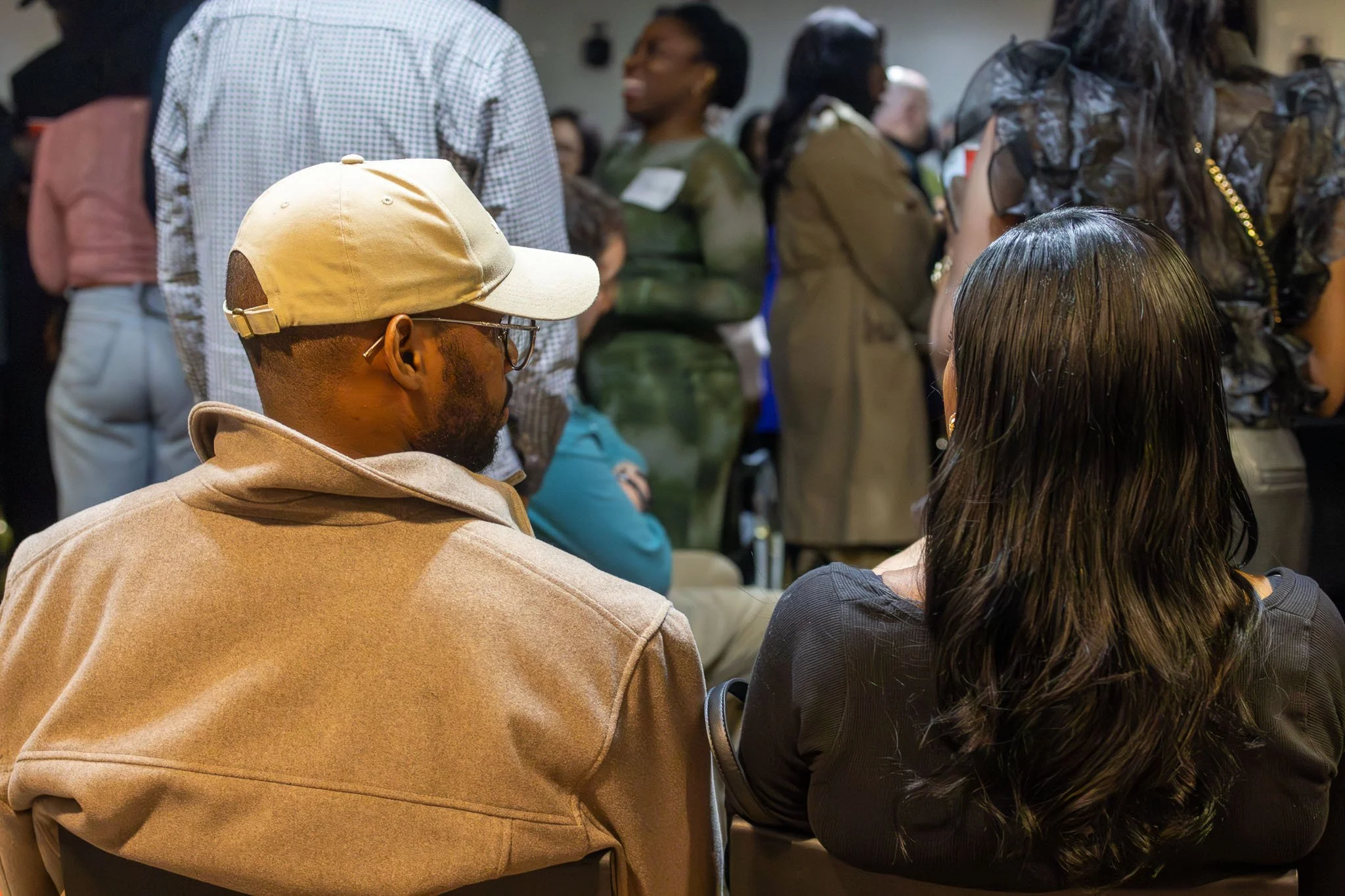 People at a crowded indoor event, sitting and standing closely together, engaging in conversations, with some individuals wearing glasses and a variety of casual clothing.